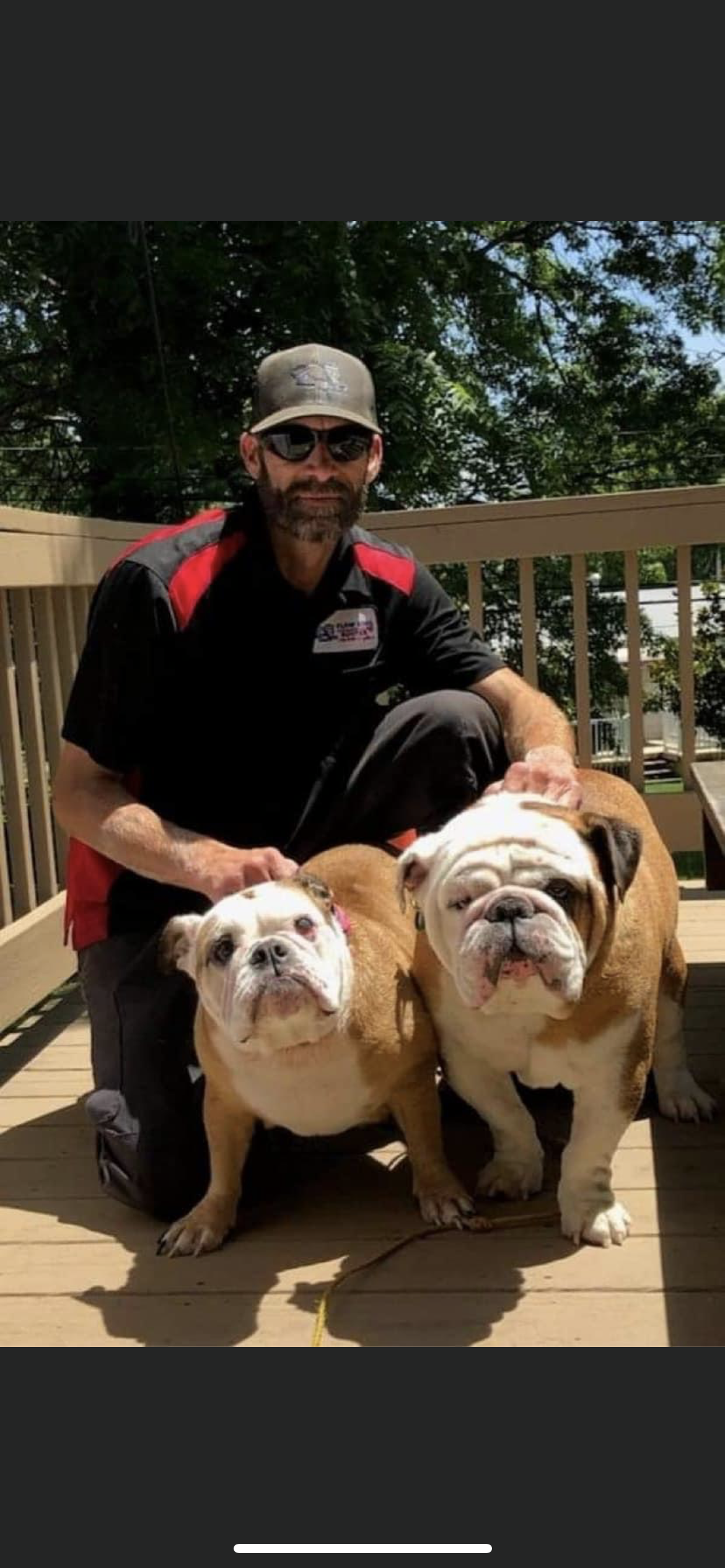 A man kneels on a deck, with two bulldogs. He wears a hat and sunglasses, holding the dogs who are brown and white.