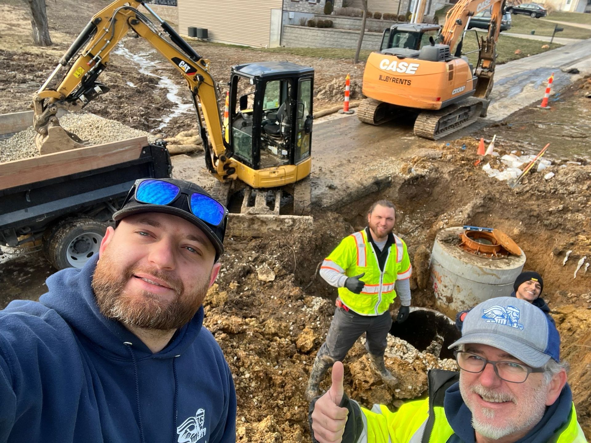 Construction workers pose for a selfie near open excavation. Excavators and dump truck are visible; one worker gives a thumbs up.