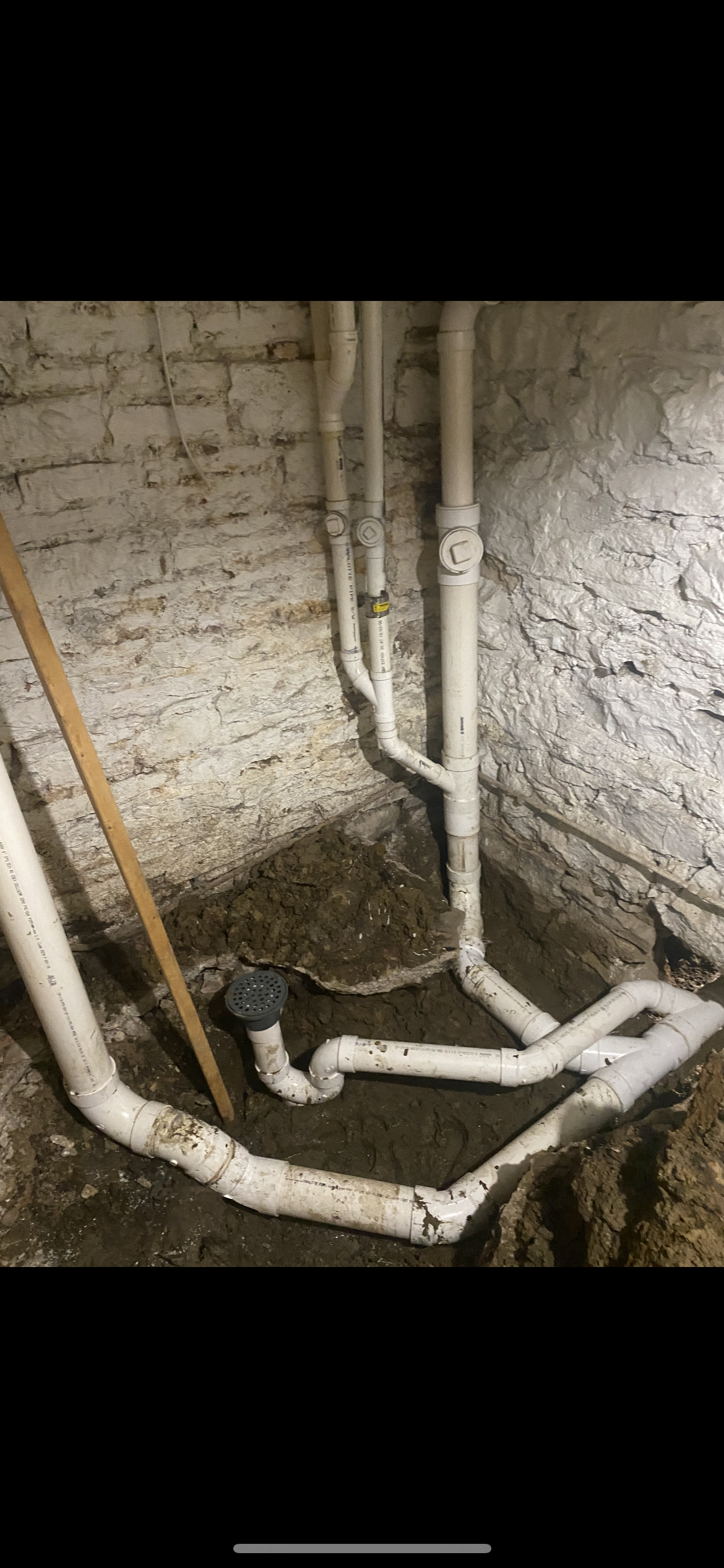 White PVC pipes in a dark basement, connected to a drain. Brick wall visible.