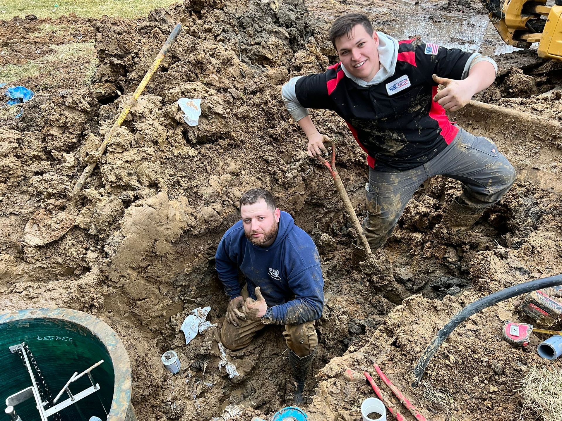 Two men in muddy clothes working on a septic system; one in hole, the other above, both giving thumbs up.