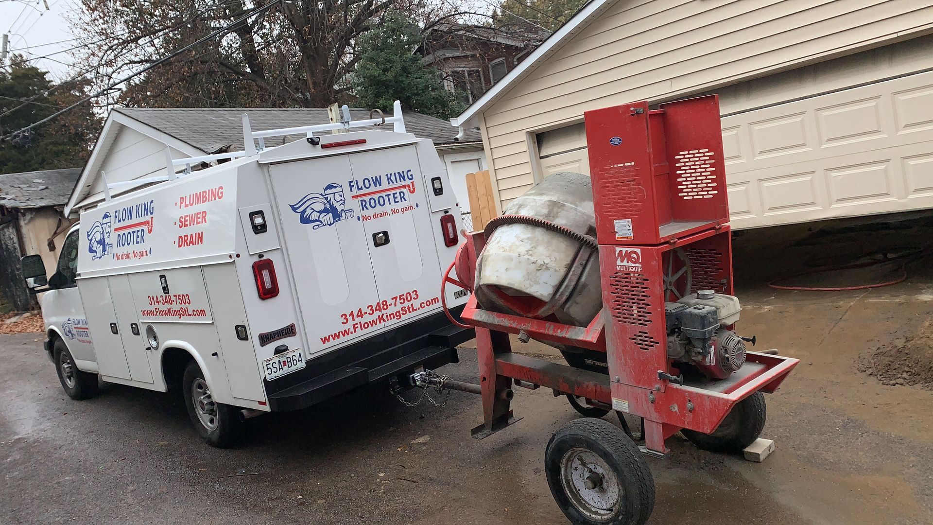 White work van parked near red cement mixer. Both are on a wet street next to a house.