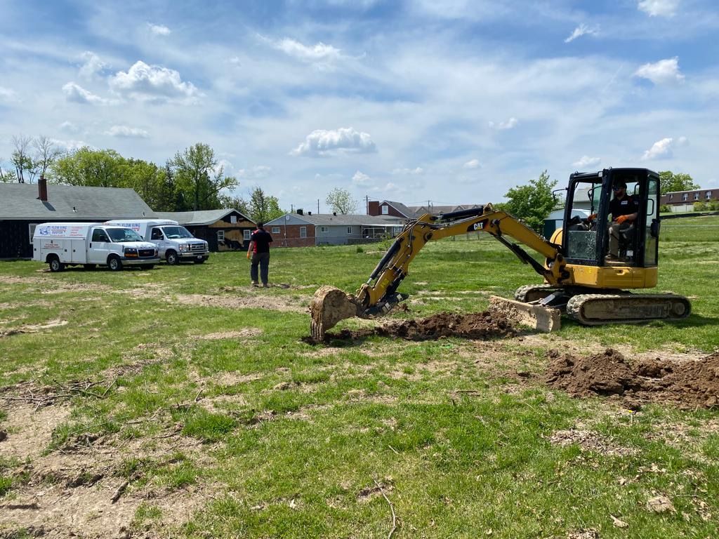 An excavator digs in a grassy field. A person stands nearby, two white vans are parked behind. Cloudy sky.