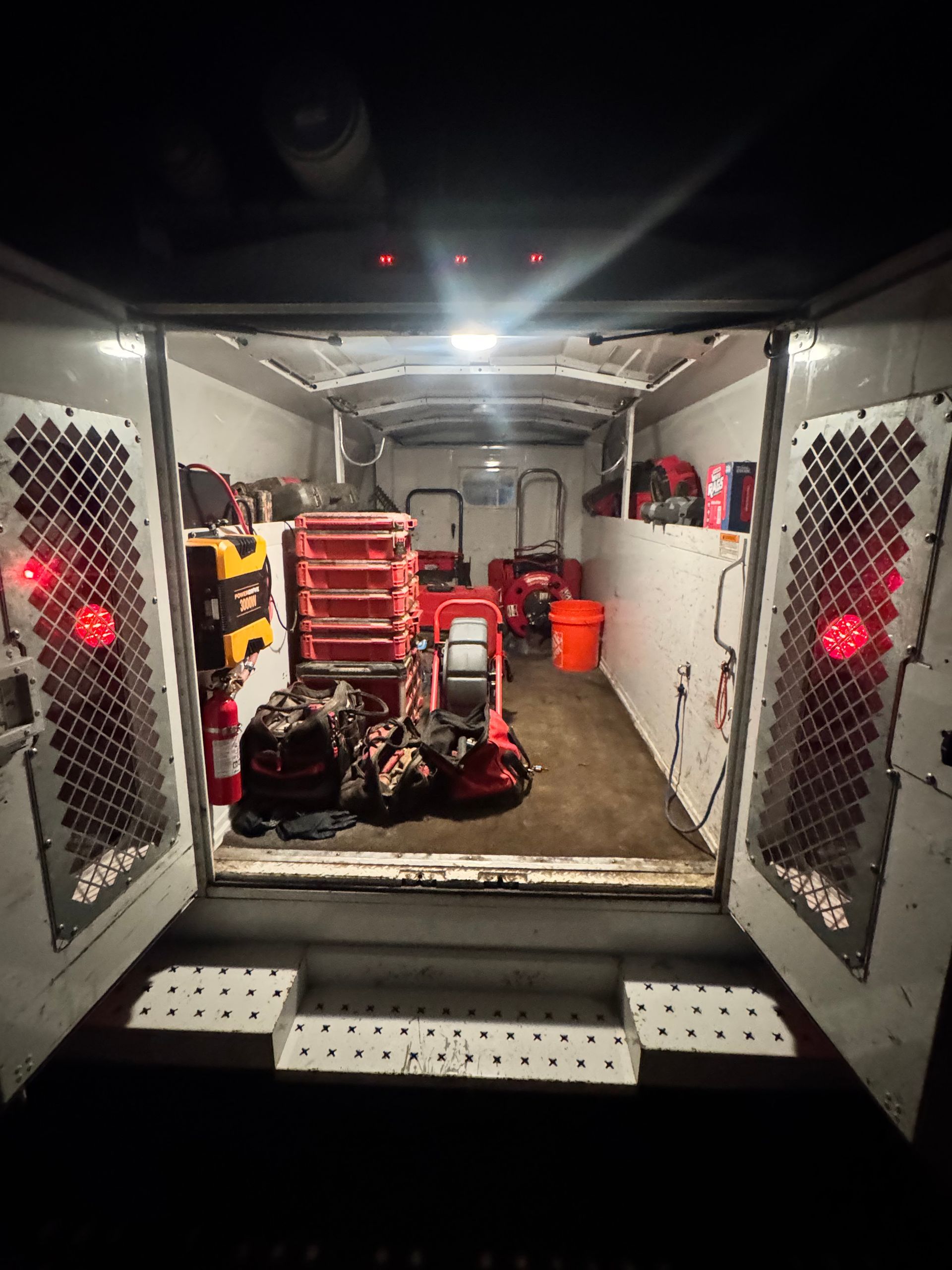 Interior of a utility trailer with tools and equipment lit by overhead lights. Open doors reveal a loading area.