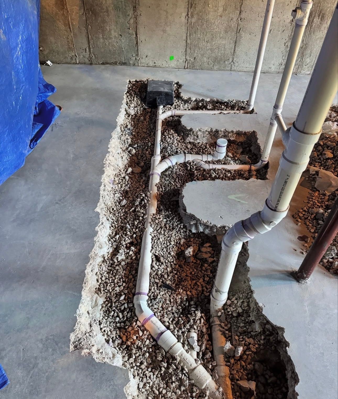 Pipes in a trench dug in a concrete floor. Gray and white pipes are connected to a square, black drain.