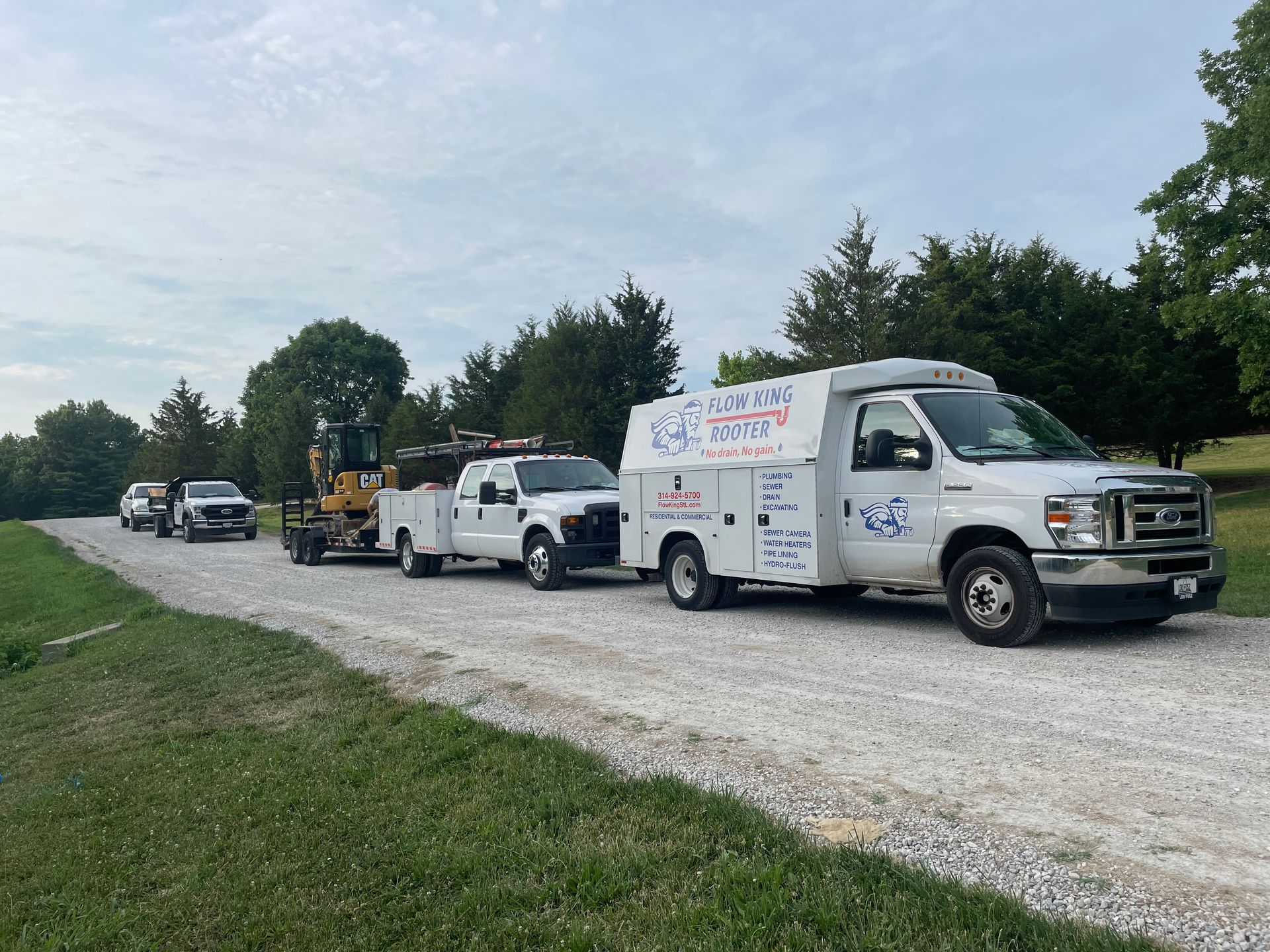 A convoy of service trucks, including one with company logos, on a gravel road, near greenery.