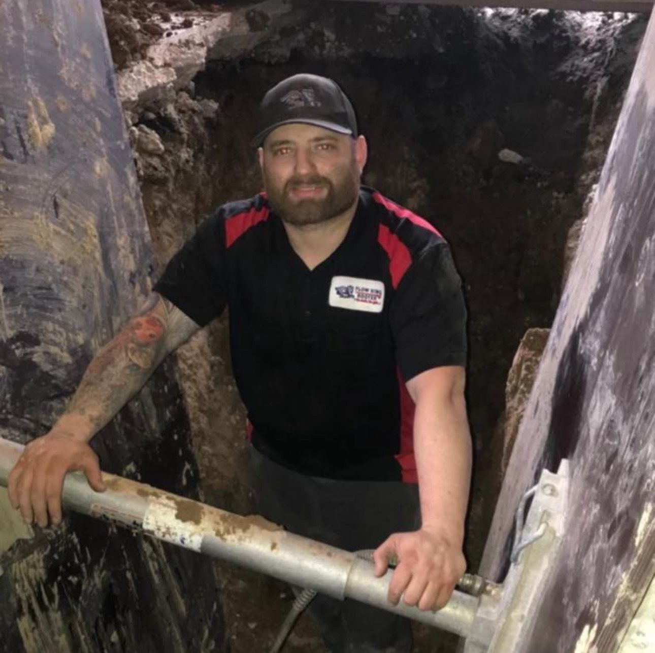 Man in black shirt, cap, in a trench, holding onto metal pipe.