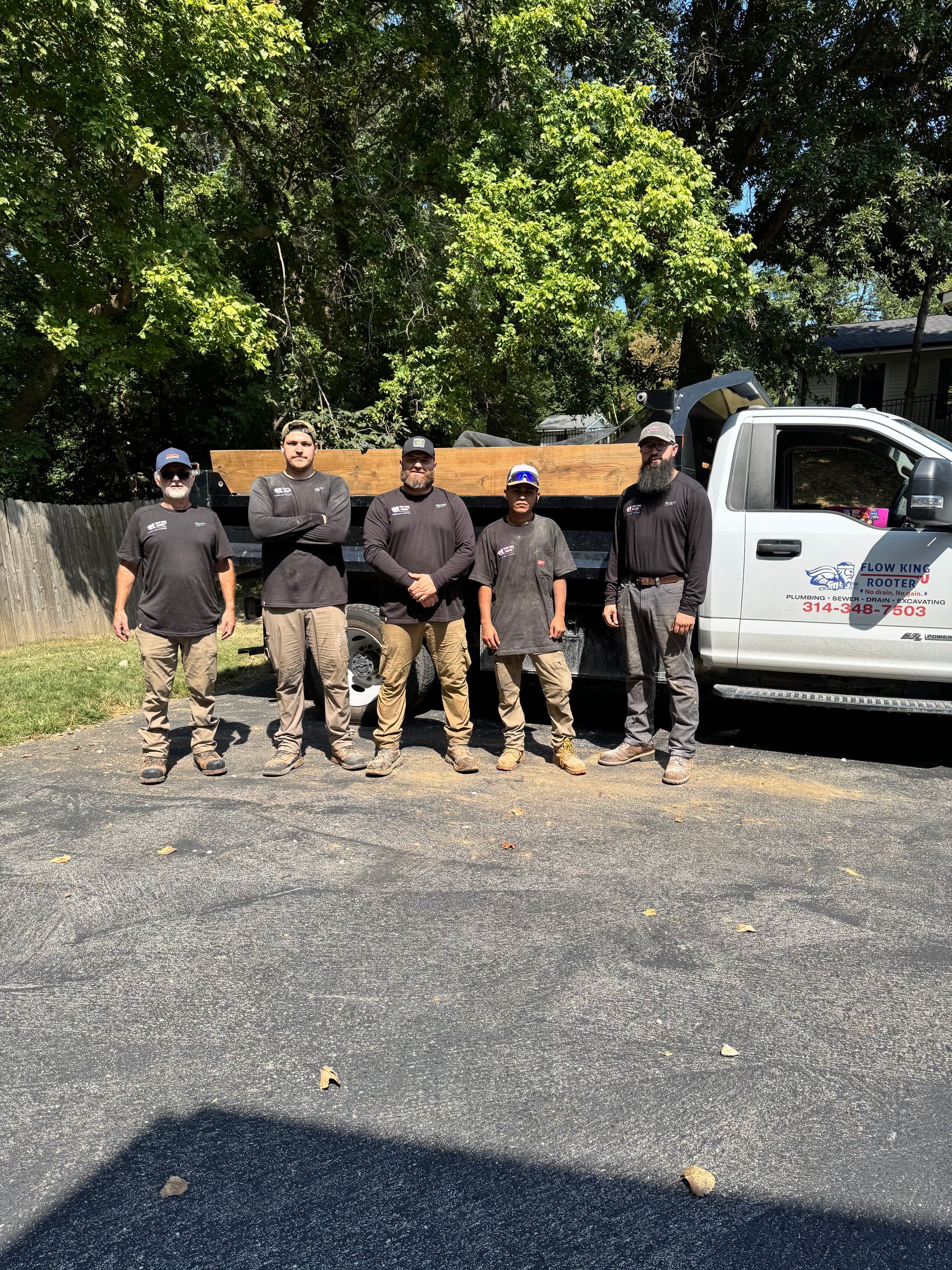 Five people pose next to a truck with a wooden beam. They wear work clothes and stand in a driveway on a sunny day.