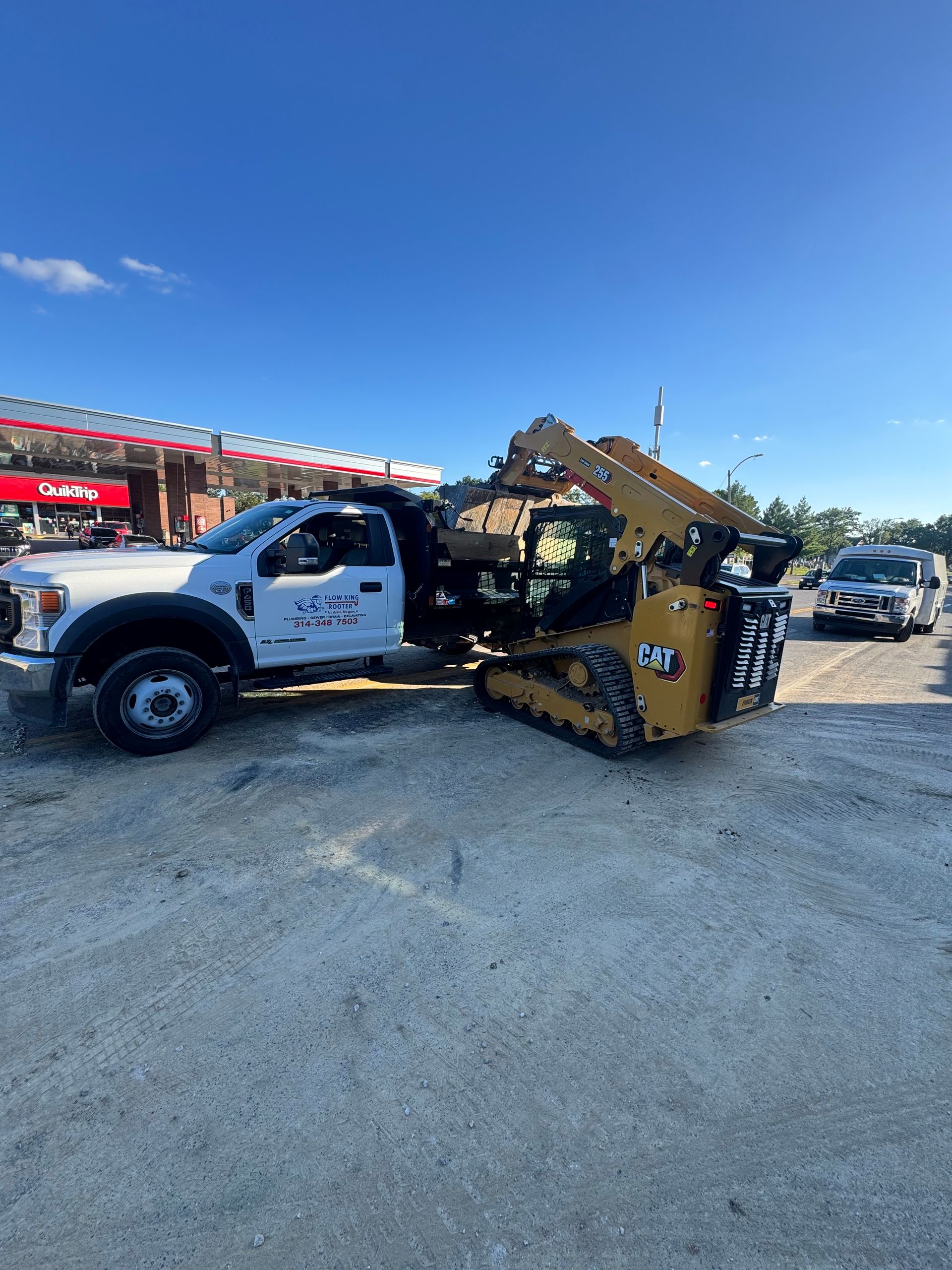 White truck with a skid steer on a trailer, parked outside a gas station on a sunny day.