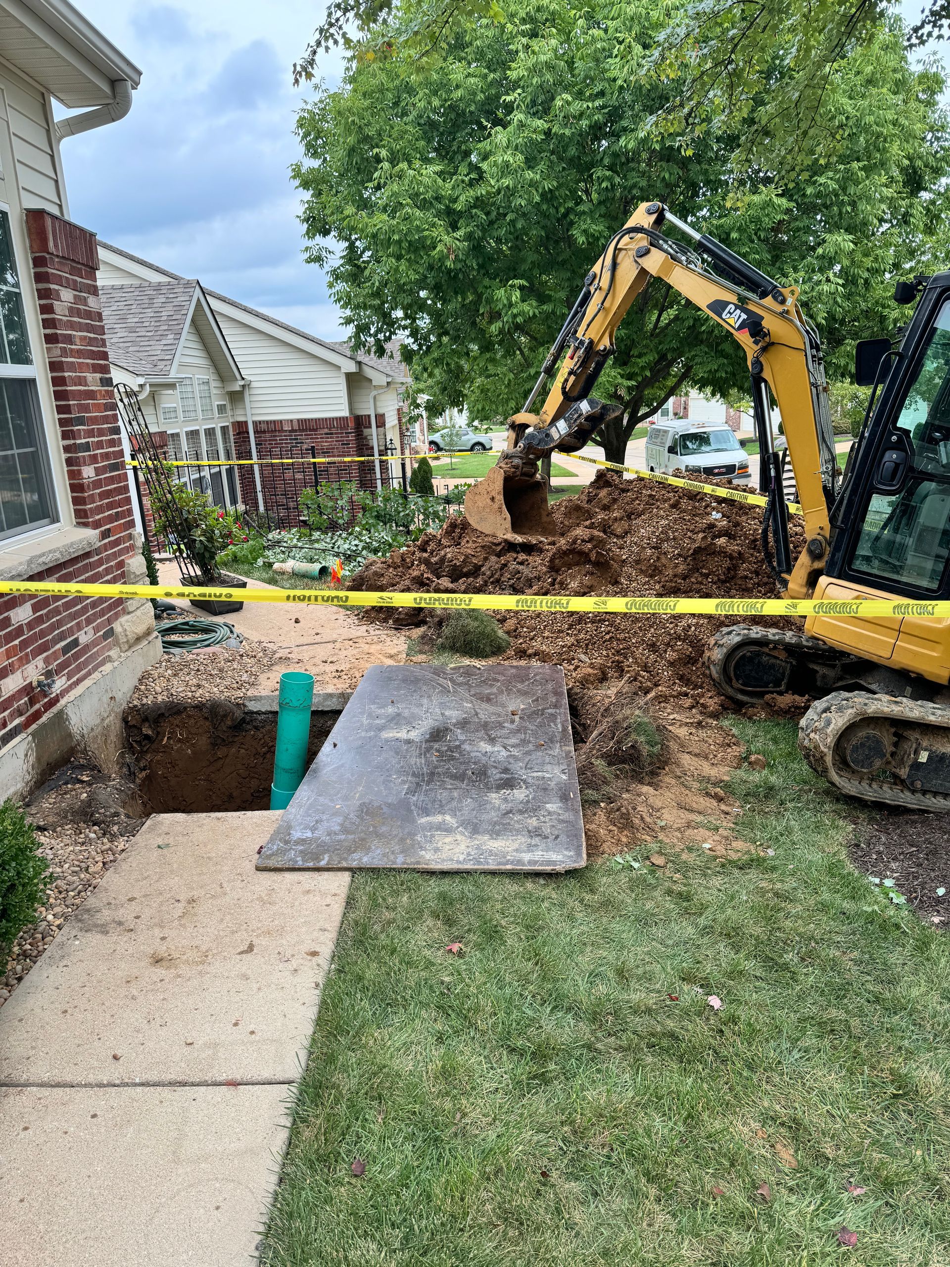 A small yellow excavator digging near a house, exposing a pipe. Caution tape surrounds the work area.