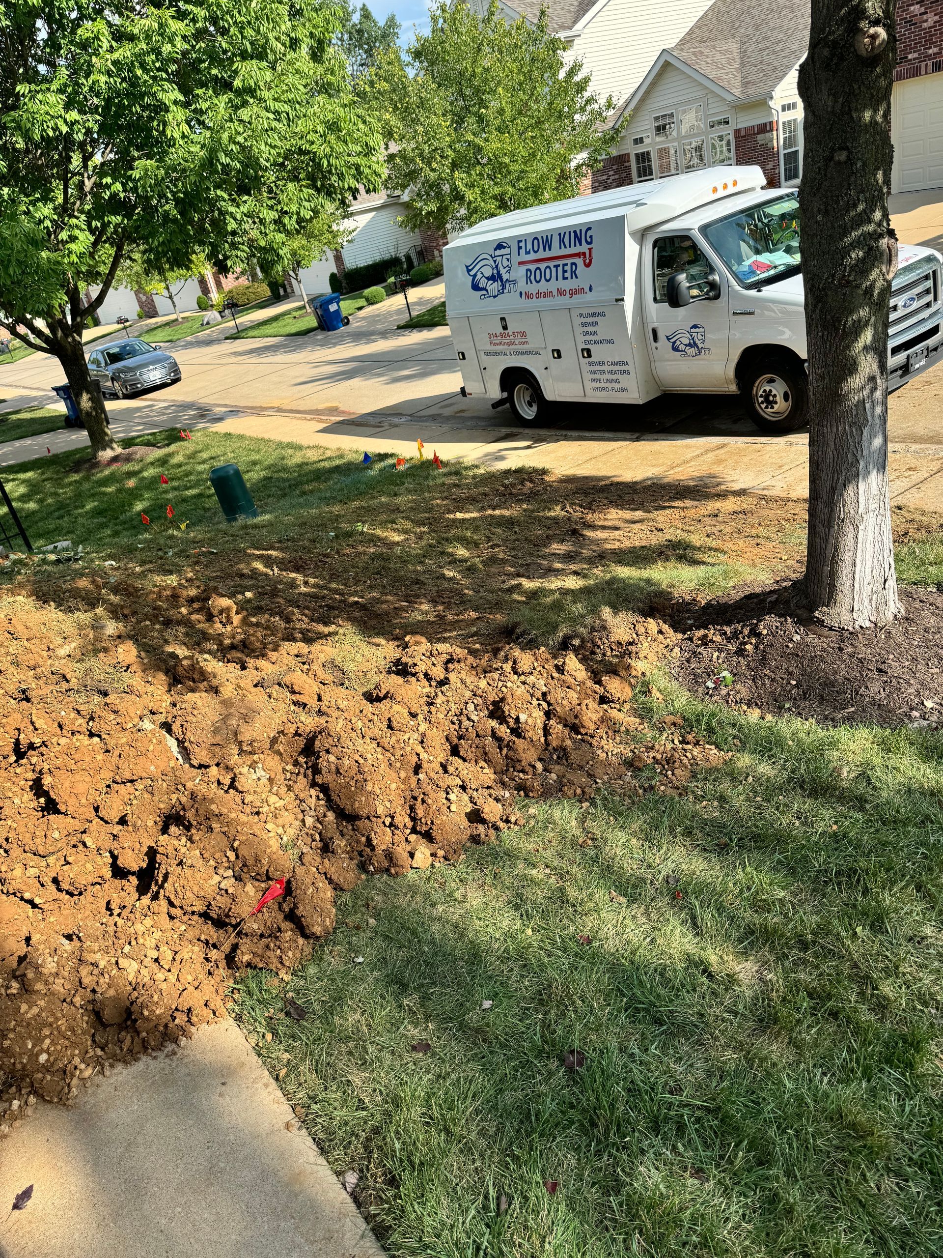 A white service van parked near a dirt pile on a lawn next to a tree and sidewalk.