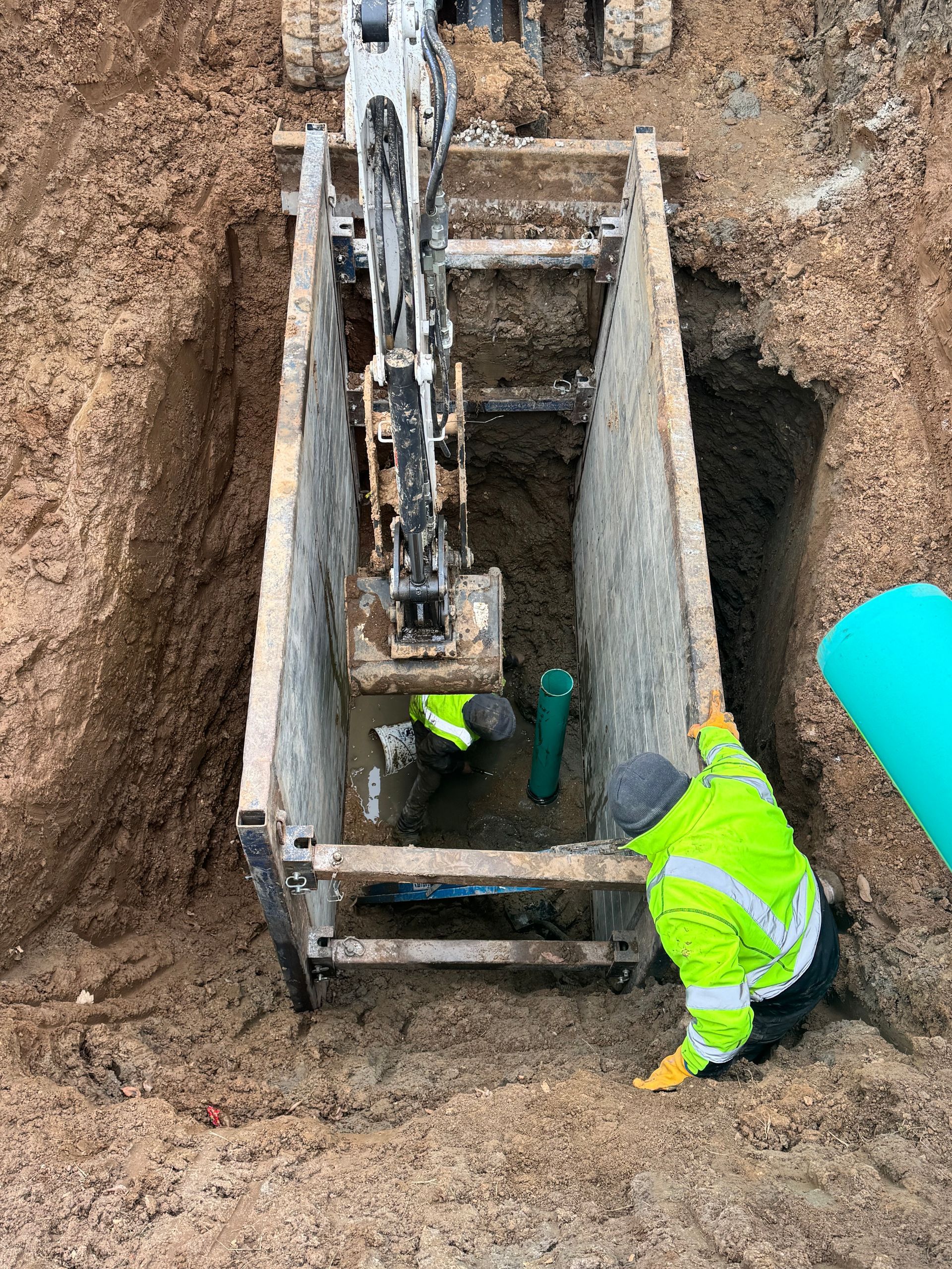 Workers in a trench installing pipe with an excavator; one in a high-vis vest.