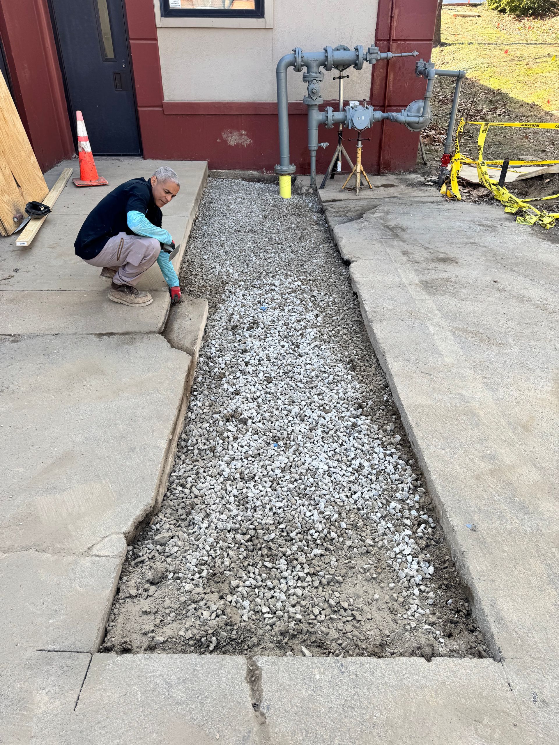 Person working on a concrete sidewalk with gravel. Near a building with gas pipes. Construction area.