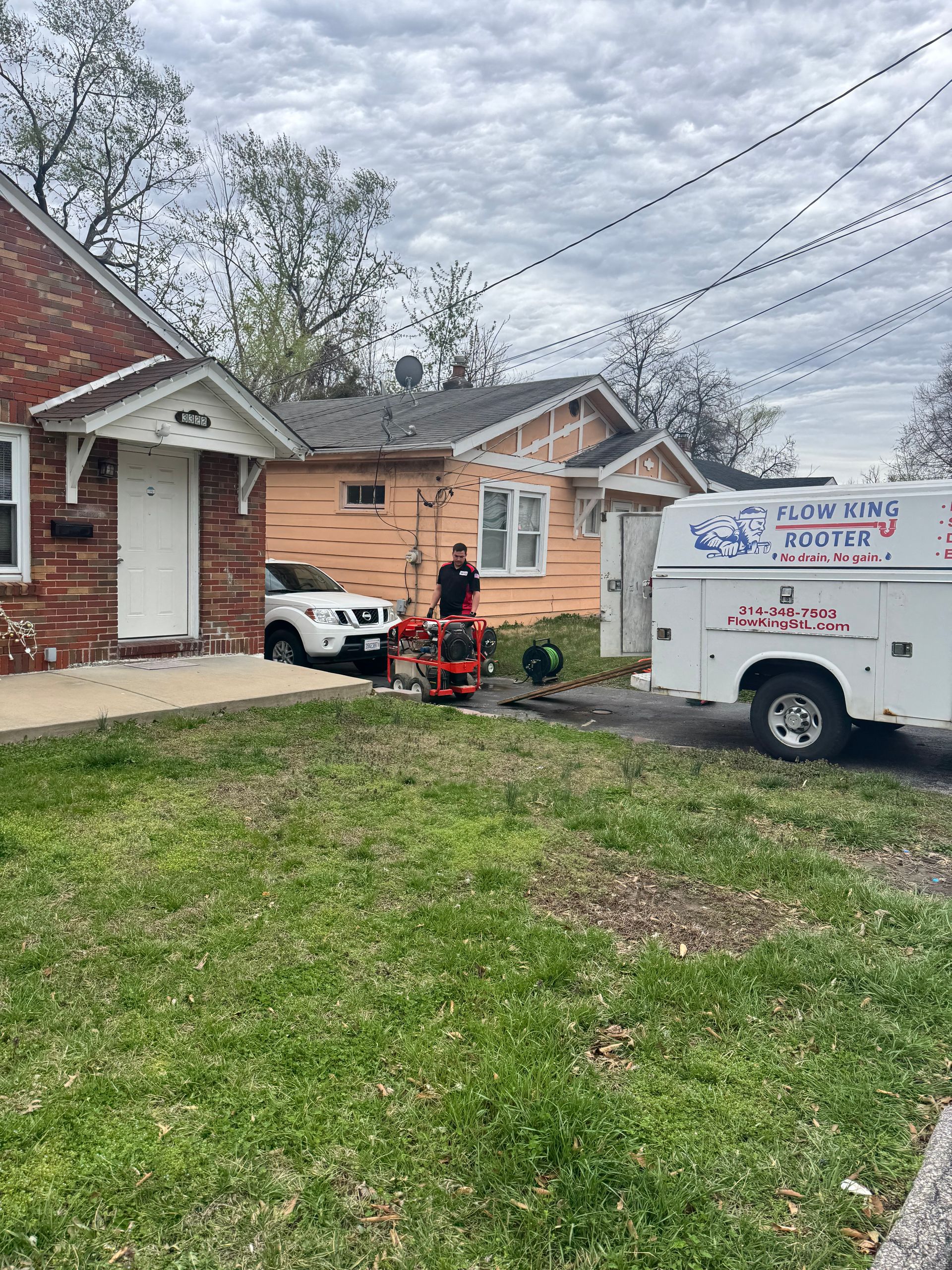Plumbing truck parked in front of homes; worker operates lift. Cloudy day.