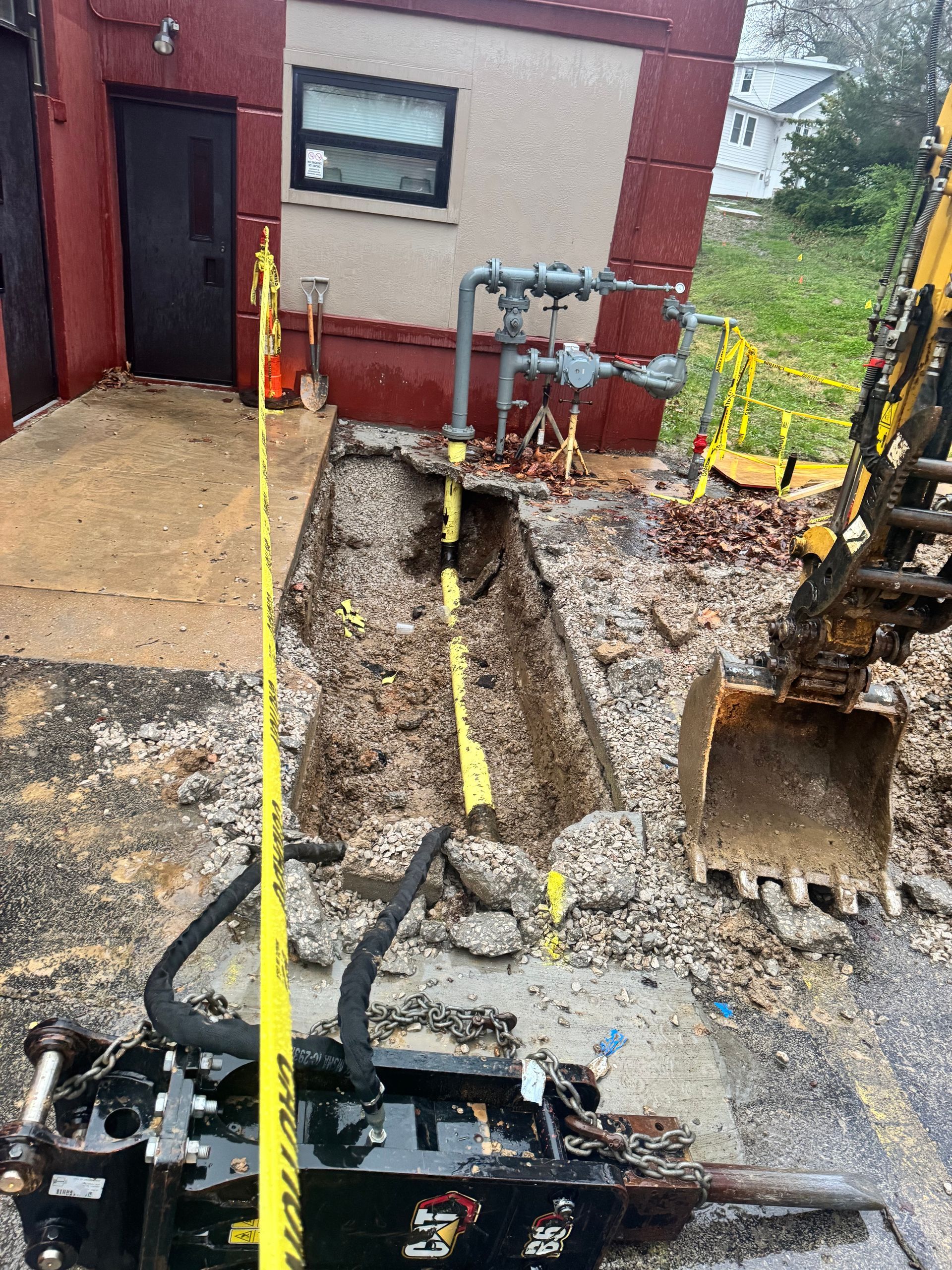 Excavation around gas lines next to a building. Yellow caution tape and an excavator are visible.