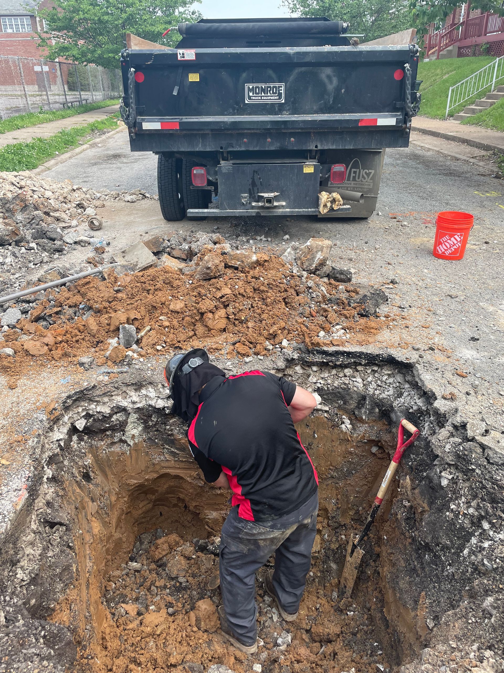 A worker digs in a square hole in a street, a dump truck is parked nearby.