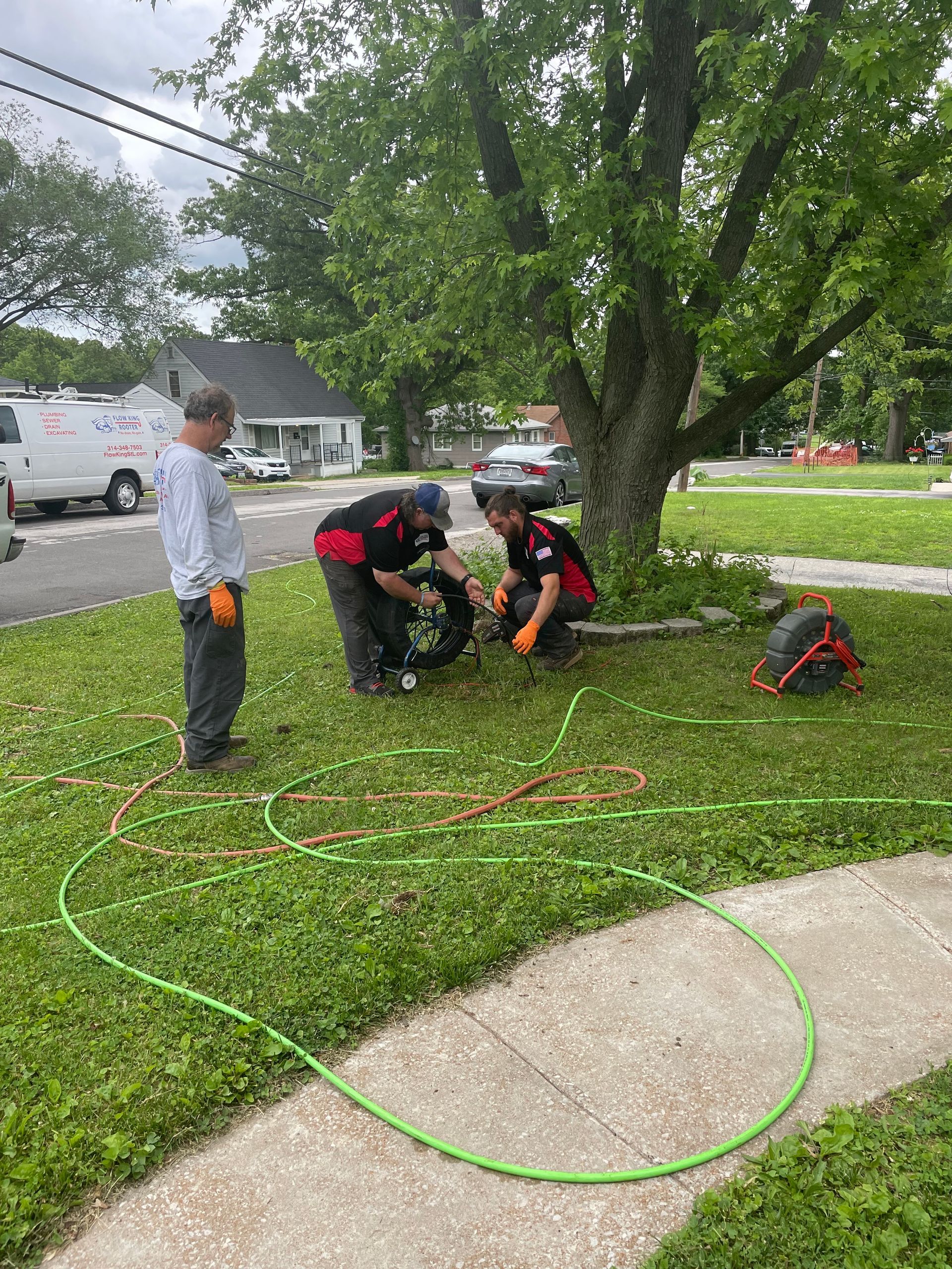 Three people working on plumbing on a lawn; one watches, two work at a pipe. Green hose sprawls on the grass.