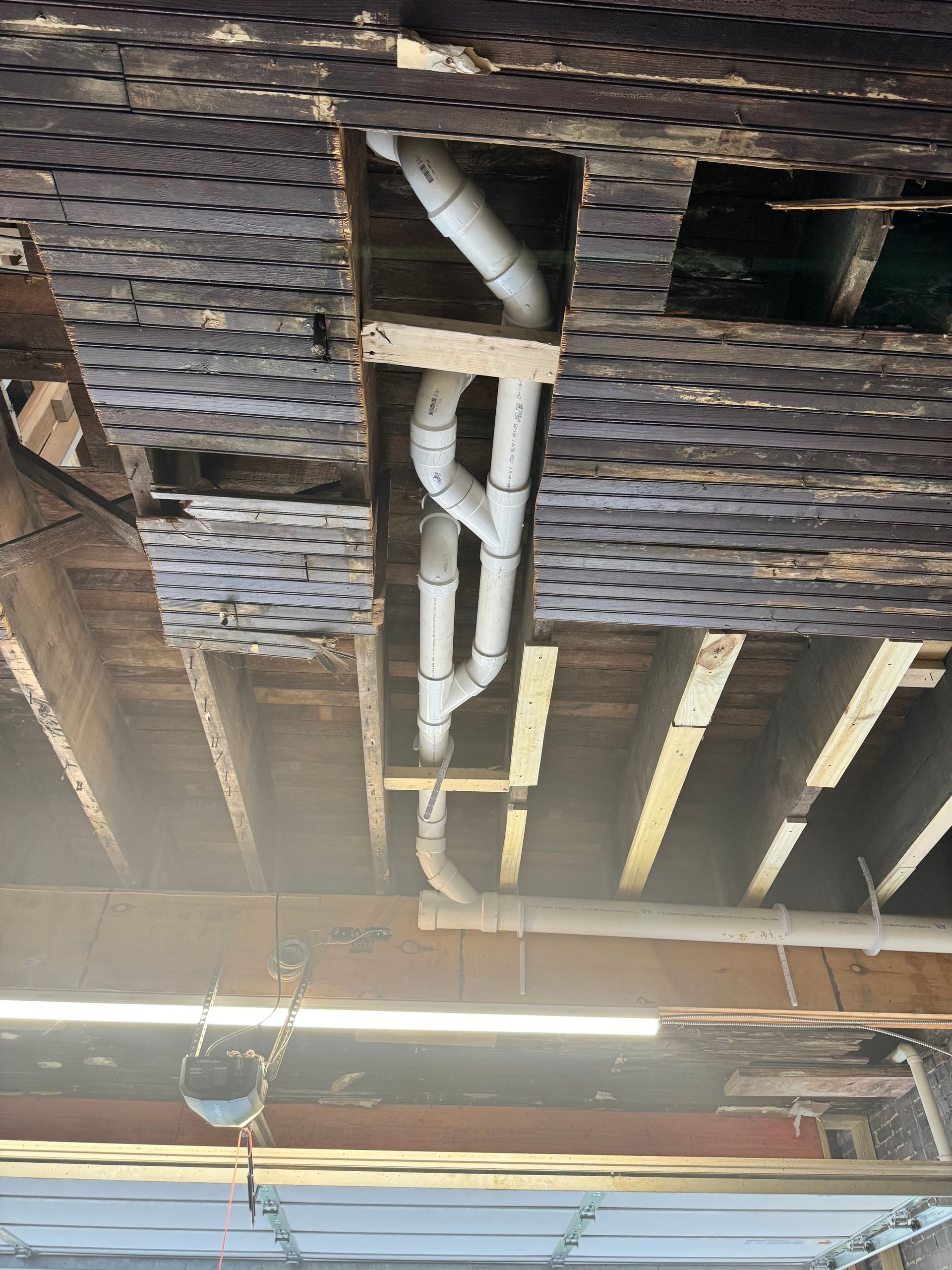 White PVC pipes run through wooden beams and a damaged, grated ceiling in a building's interior.