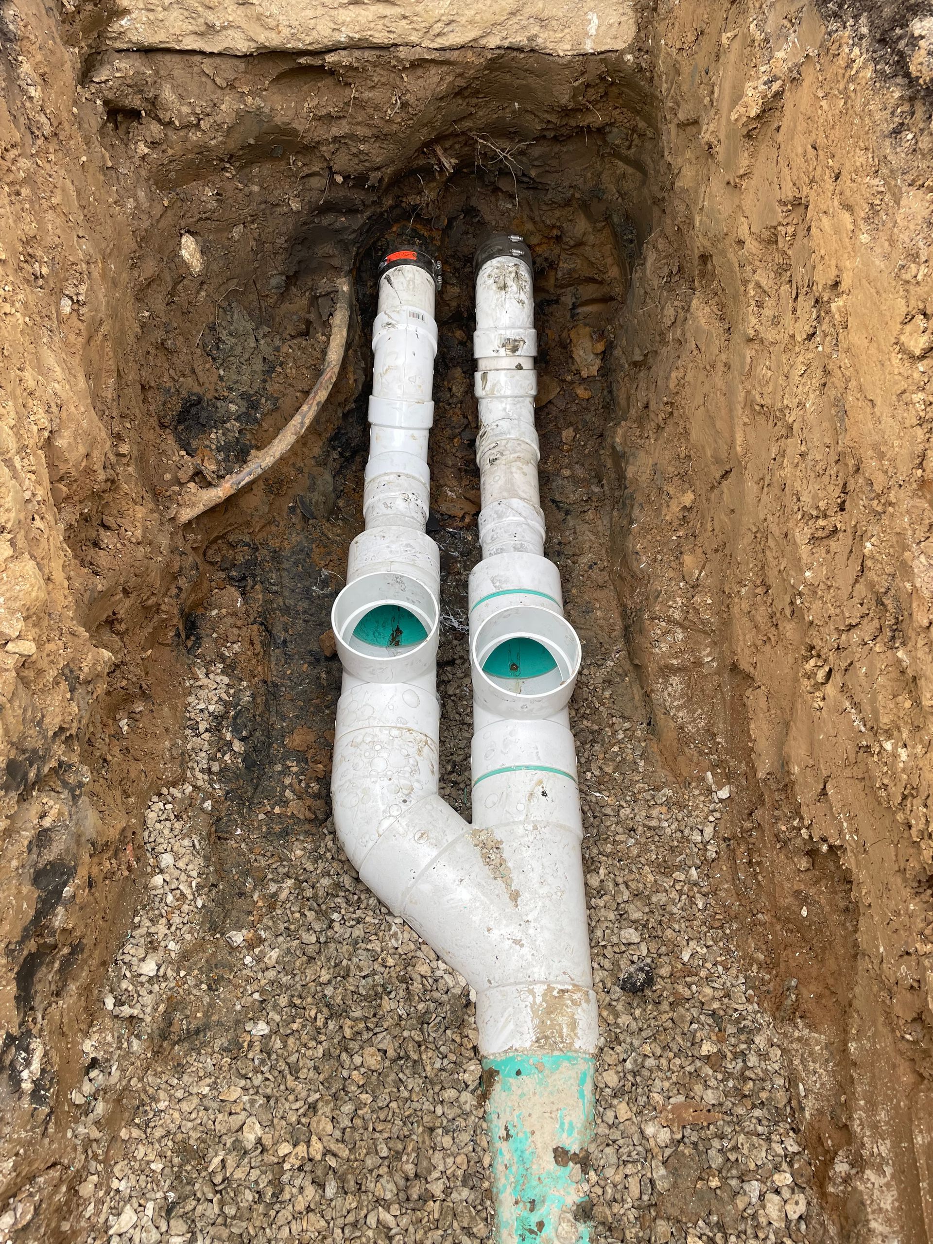 PVC pipes in an excavated trench. Two vertical pipes connect to a single, lower pipe with a Y-shaped fitting.