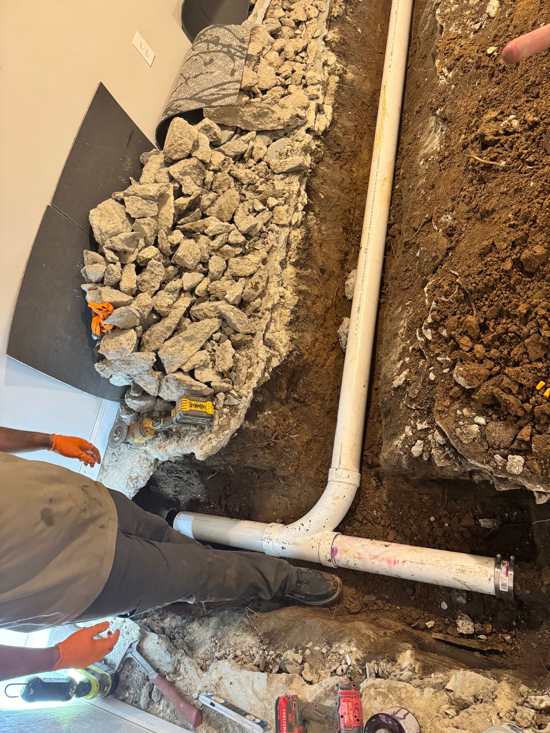 Plumbing work in progress: white PVC pipes laid in a trench alongside gravel and soil. A person in orange gloves is present.