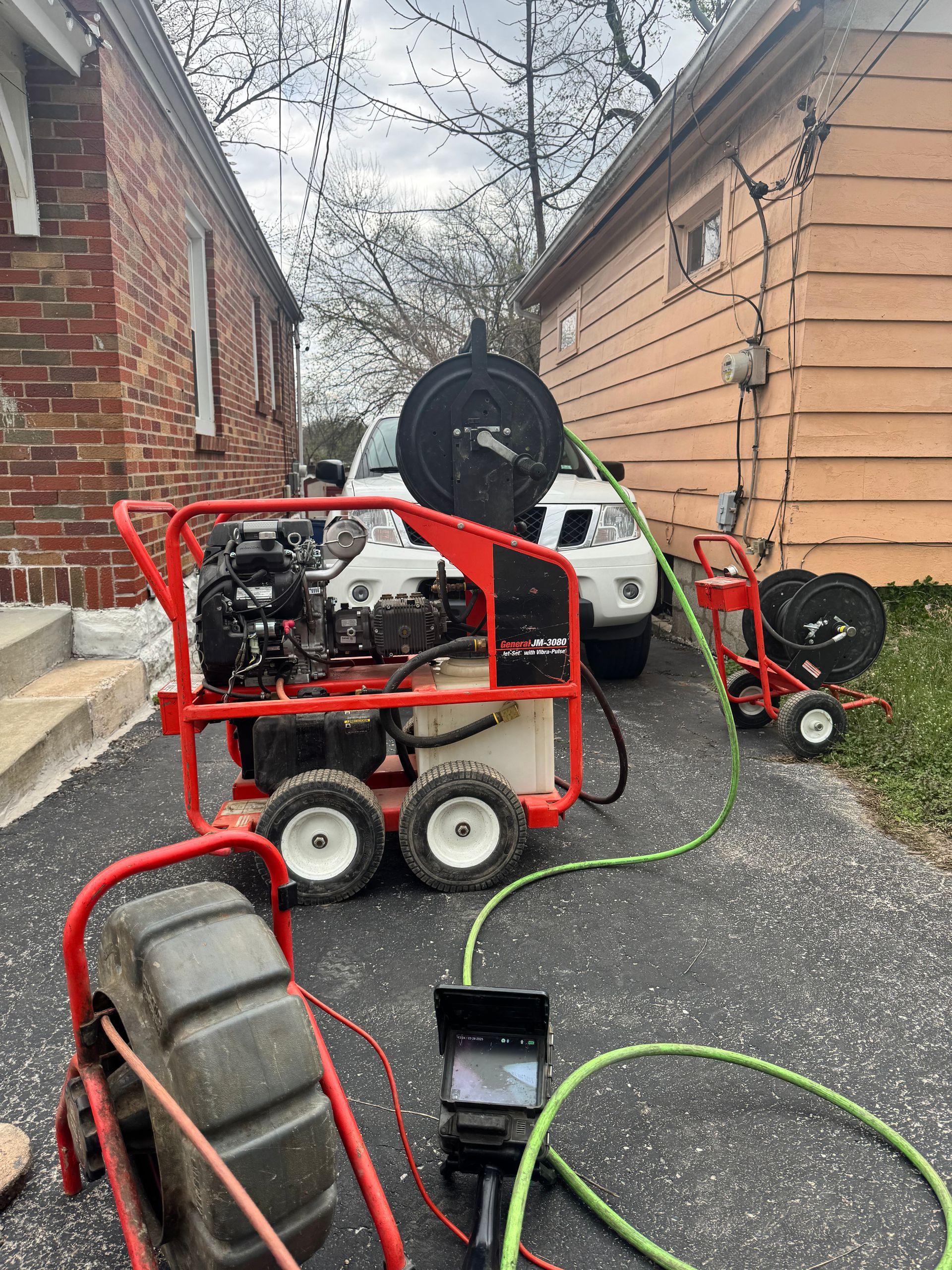 Plumbing equipment on a driveway between two houses; a sewer camera, jetter, and reel cart are connected.