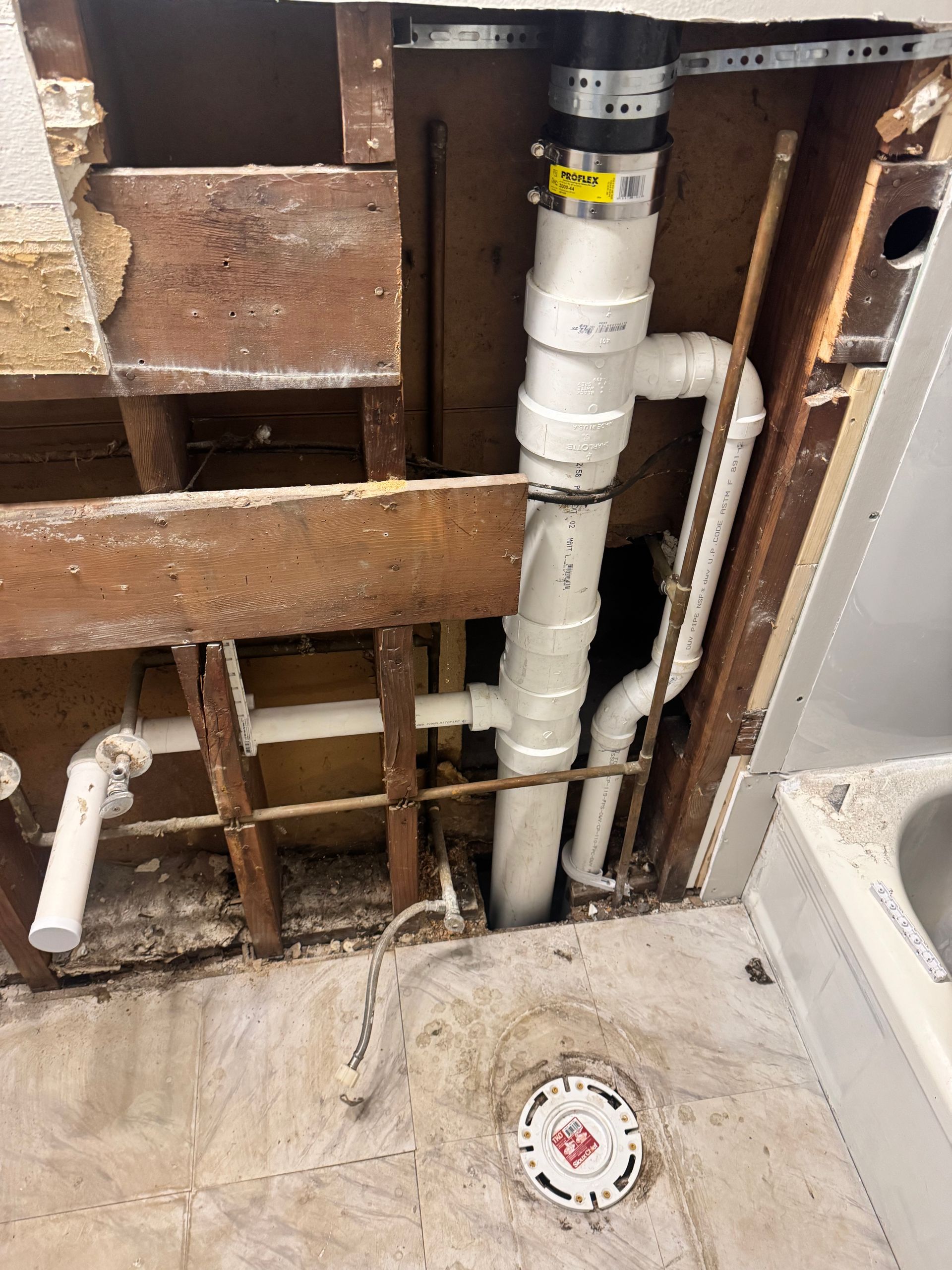 Plumbing pipes exposed in a wall, including white PVC pipes and a black fitting. A floor drain is visible.