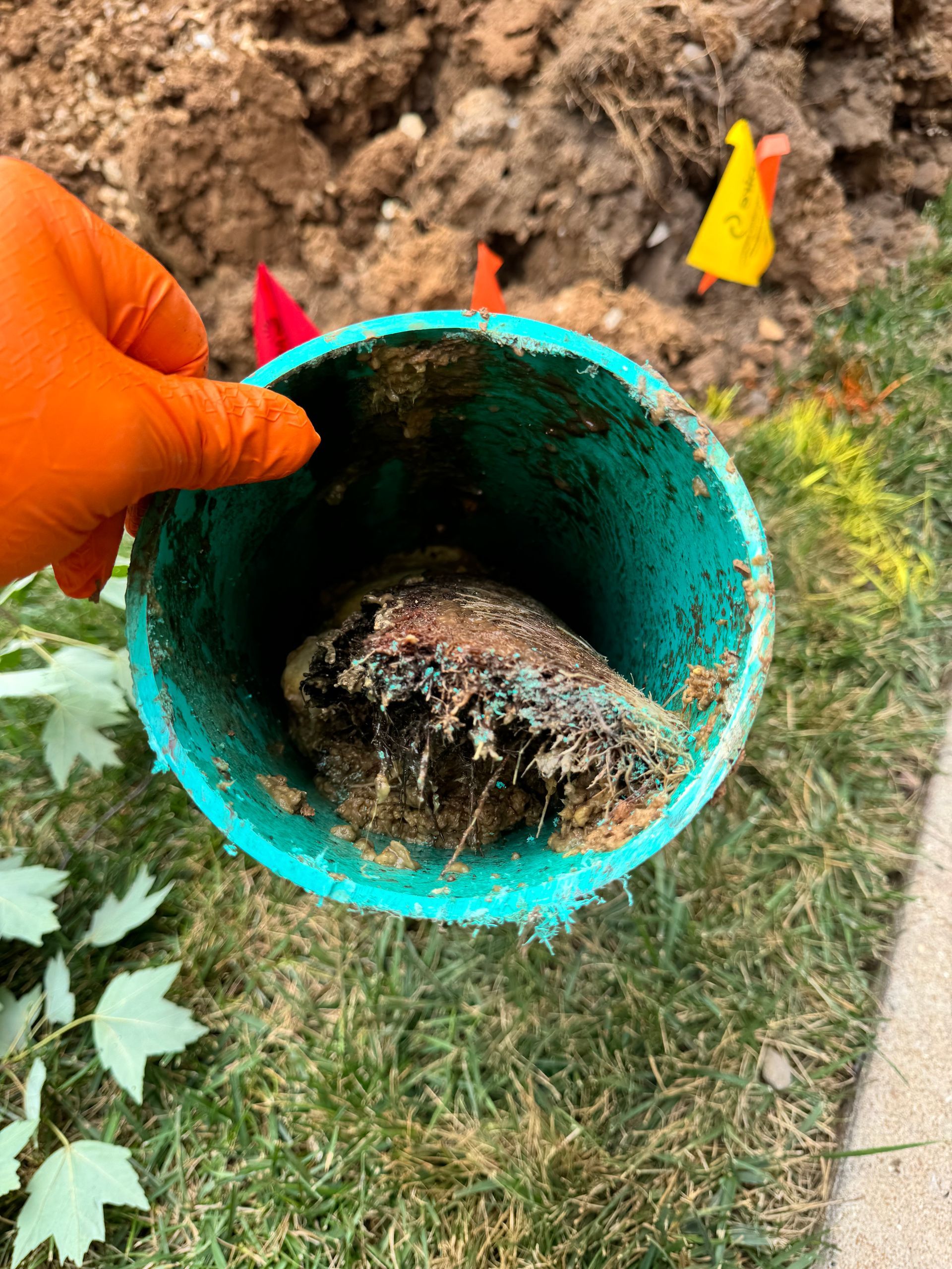 Hand holding a green pipe filled with a dark, fibrous clog, outdoors in dirt and grass.