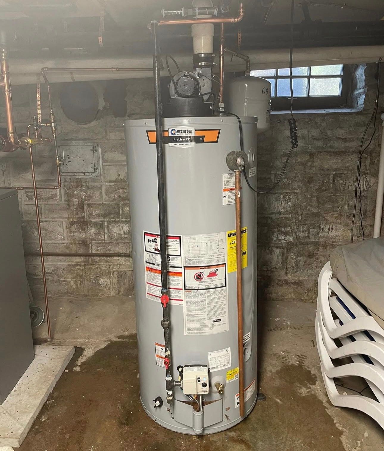 Gray water heater in a basement setting with copper pipes and a small window.