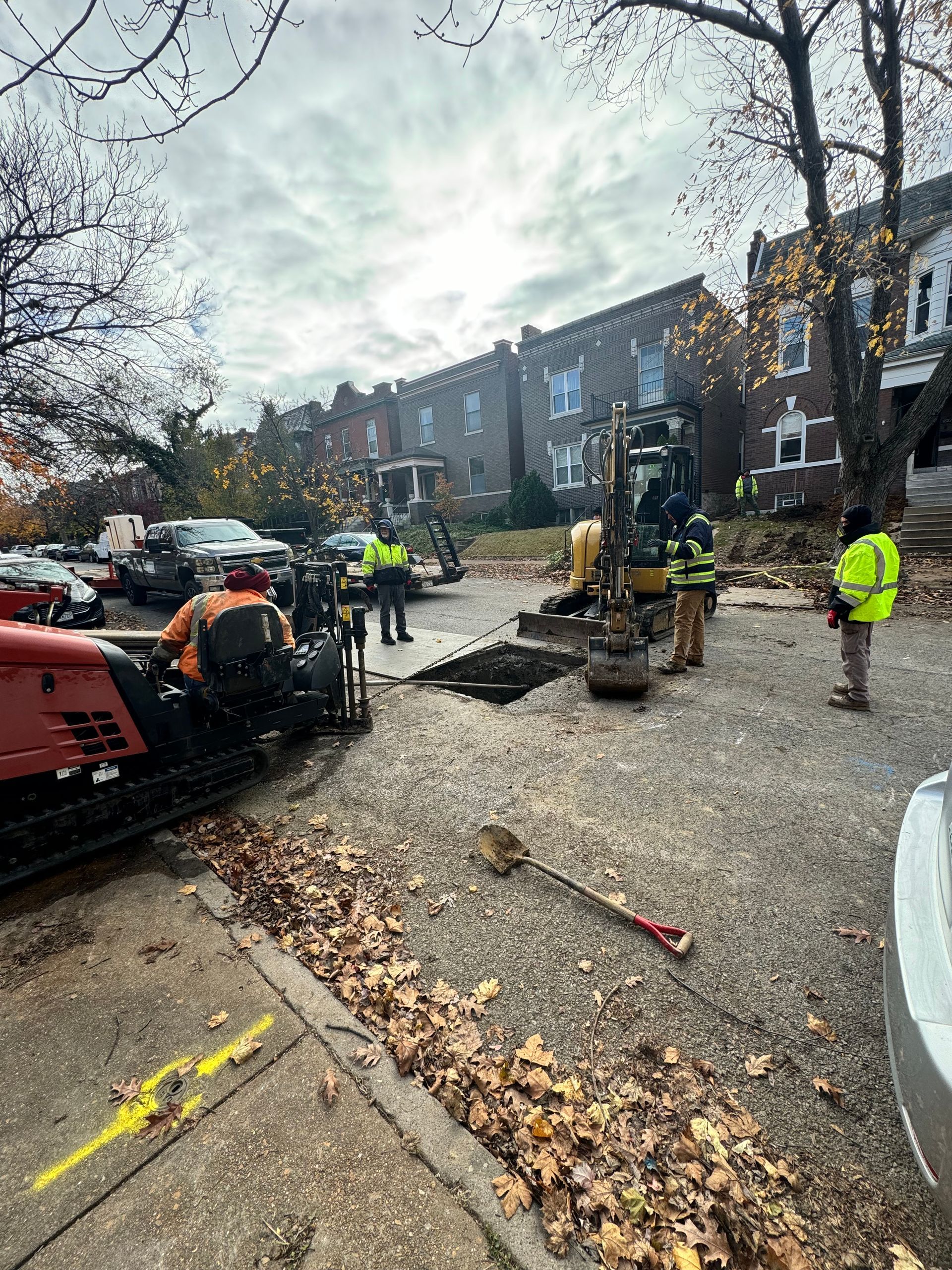 Construction workers around a hole in a gravel street; yellow safety vests, excavator, trees, brick buildings in background.