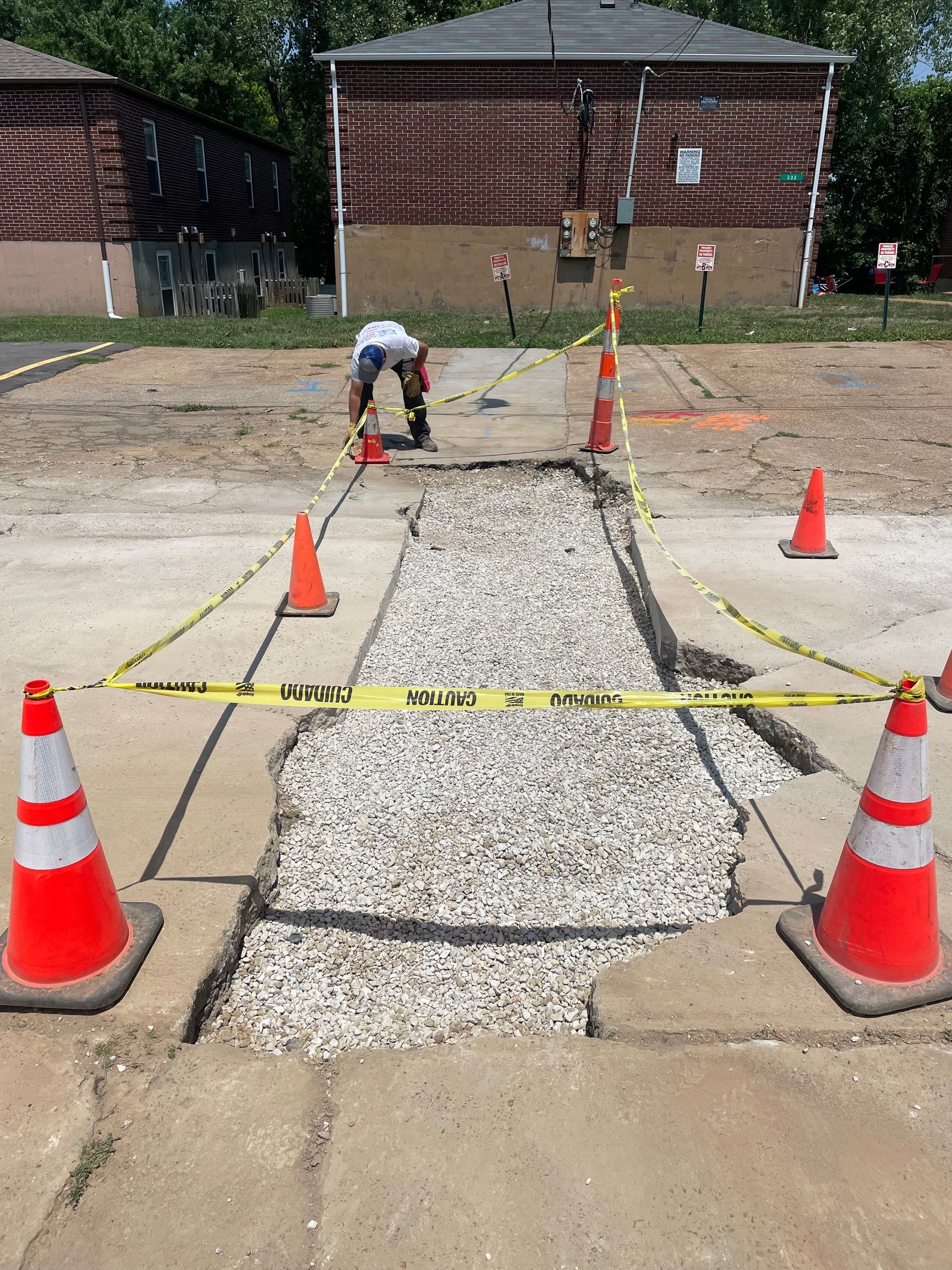 A worker repairs a sidewalk. Yellow caution tape and orange cones surround the gravel-filled area.