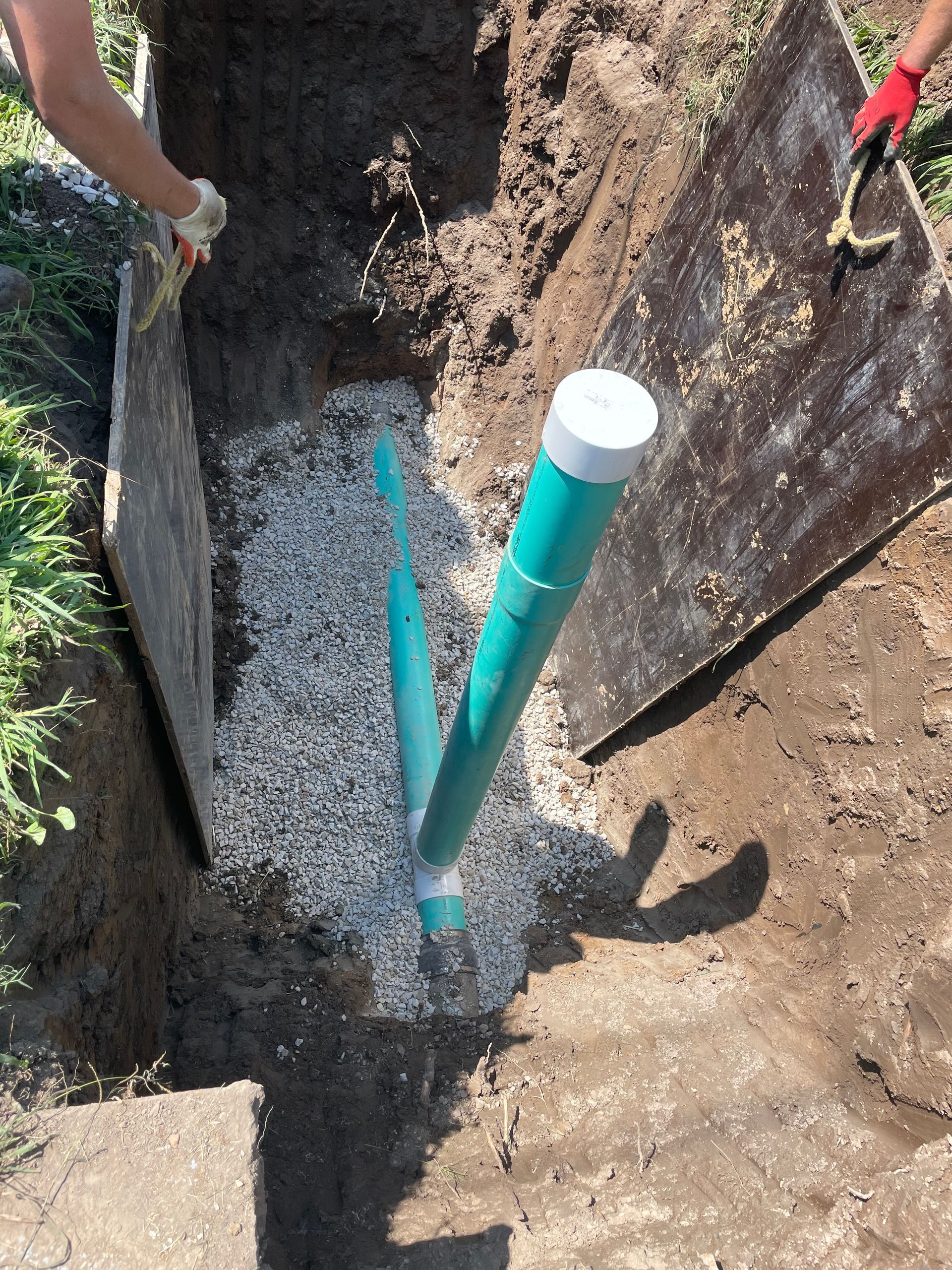 Workers installing green pipes in a trench filled with gravel. Two people hold wooden boards.
