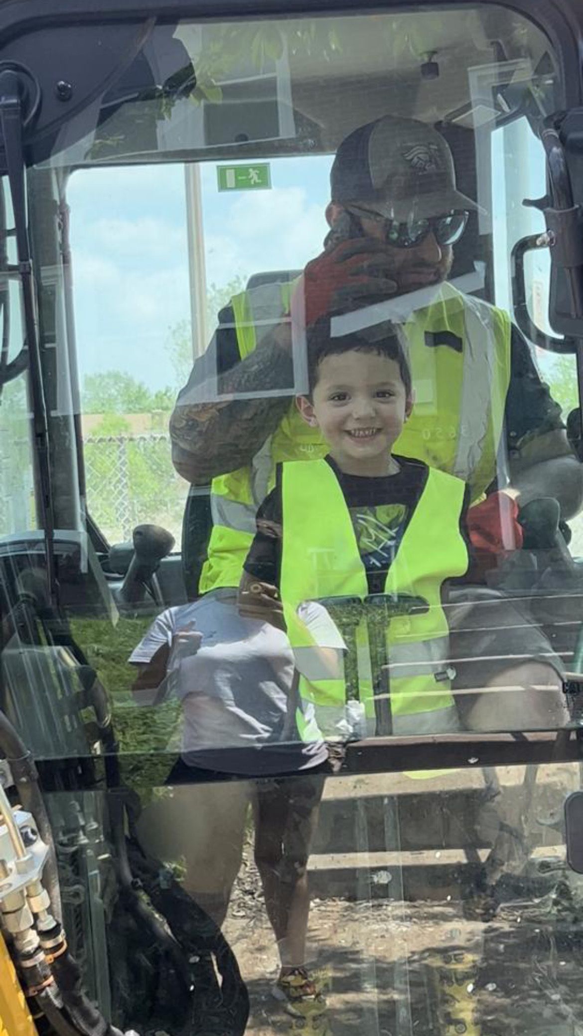 Man and child in a construction vehicle, both wearing safety vests. Child smiles brightly.