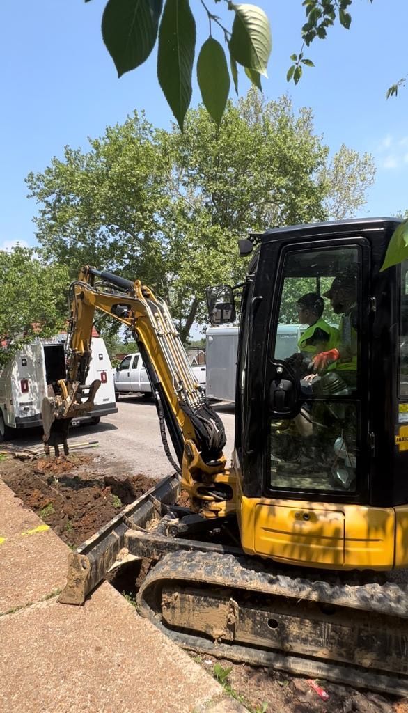 Yellow excavator digging in a dirt trench next to a sidewalk. A worker is in the cab. A white van is parked nearby.