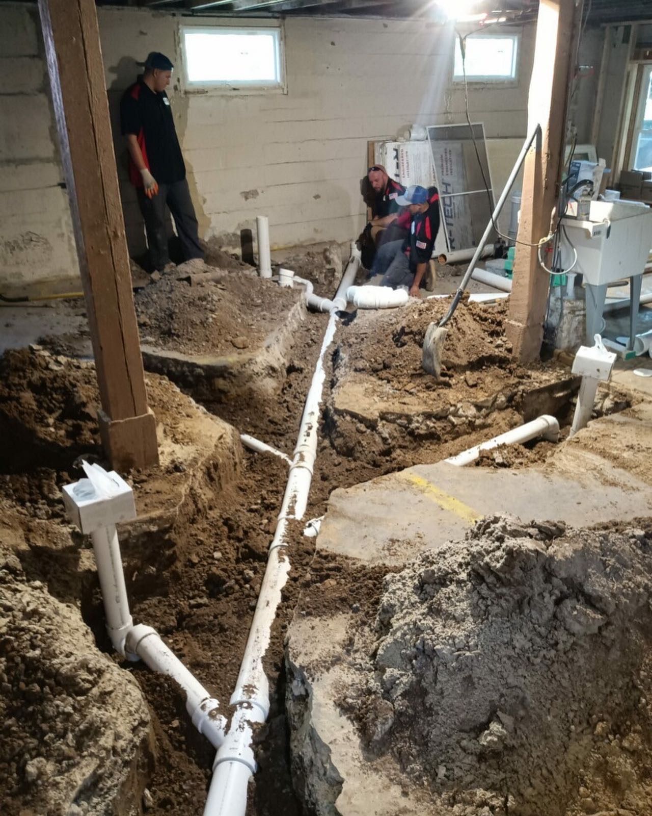 Plumbing work in a basement. Workers installing white pipes in trenches. Dirt and tools surround them.