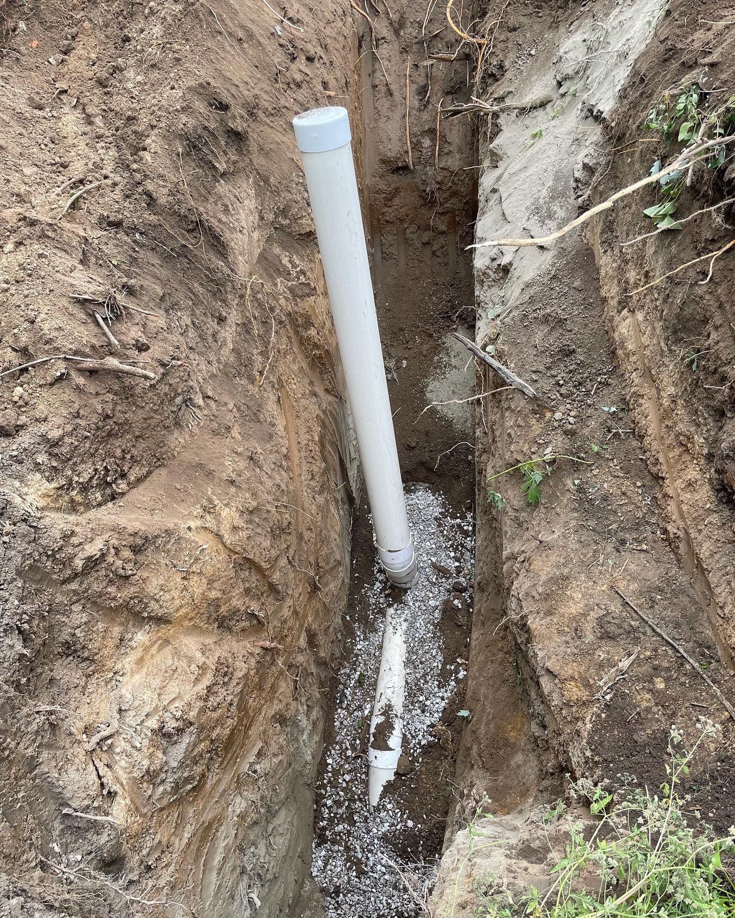 White PVC pipe in a narrow trench filled with gravel, likely for a plumbing or drainage system.