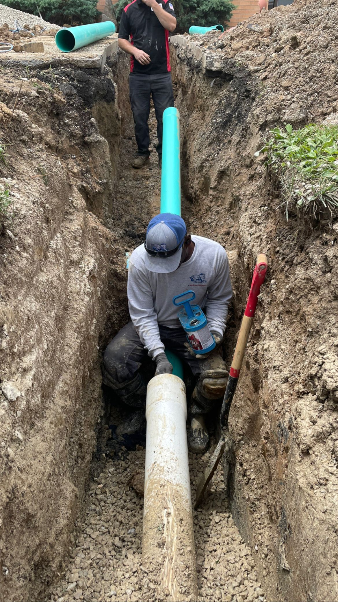 Two construction workers in a trench connecting pipes. One kneels with a tool, the other stands.