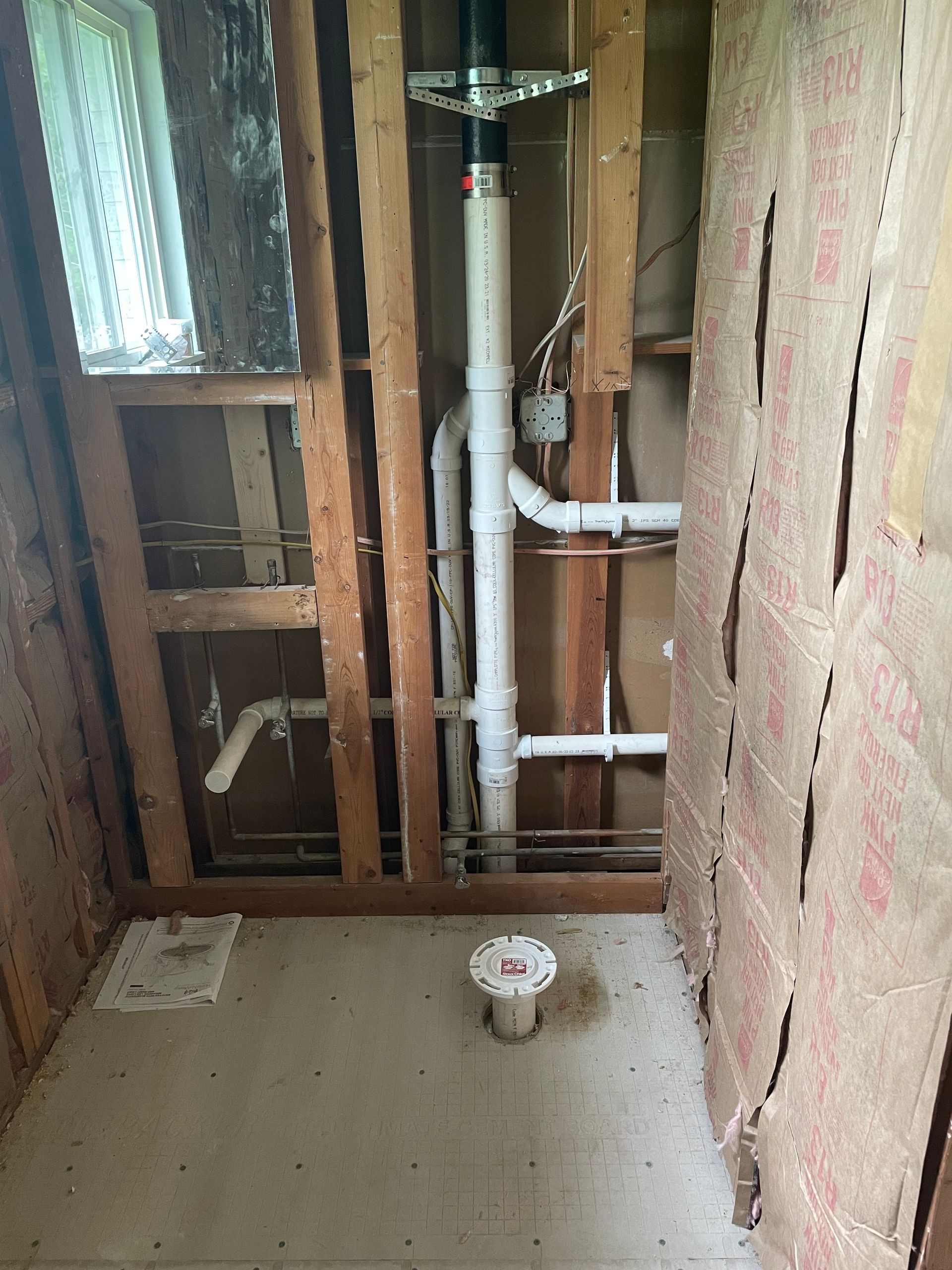 Bathroom renovation in progress, exposed plumbing and framing. White PVC pipes, floor drain, exposed insulation.