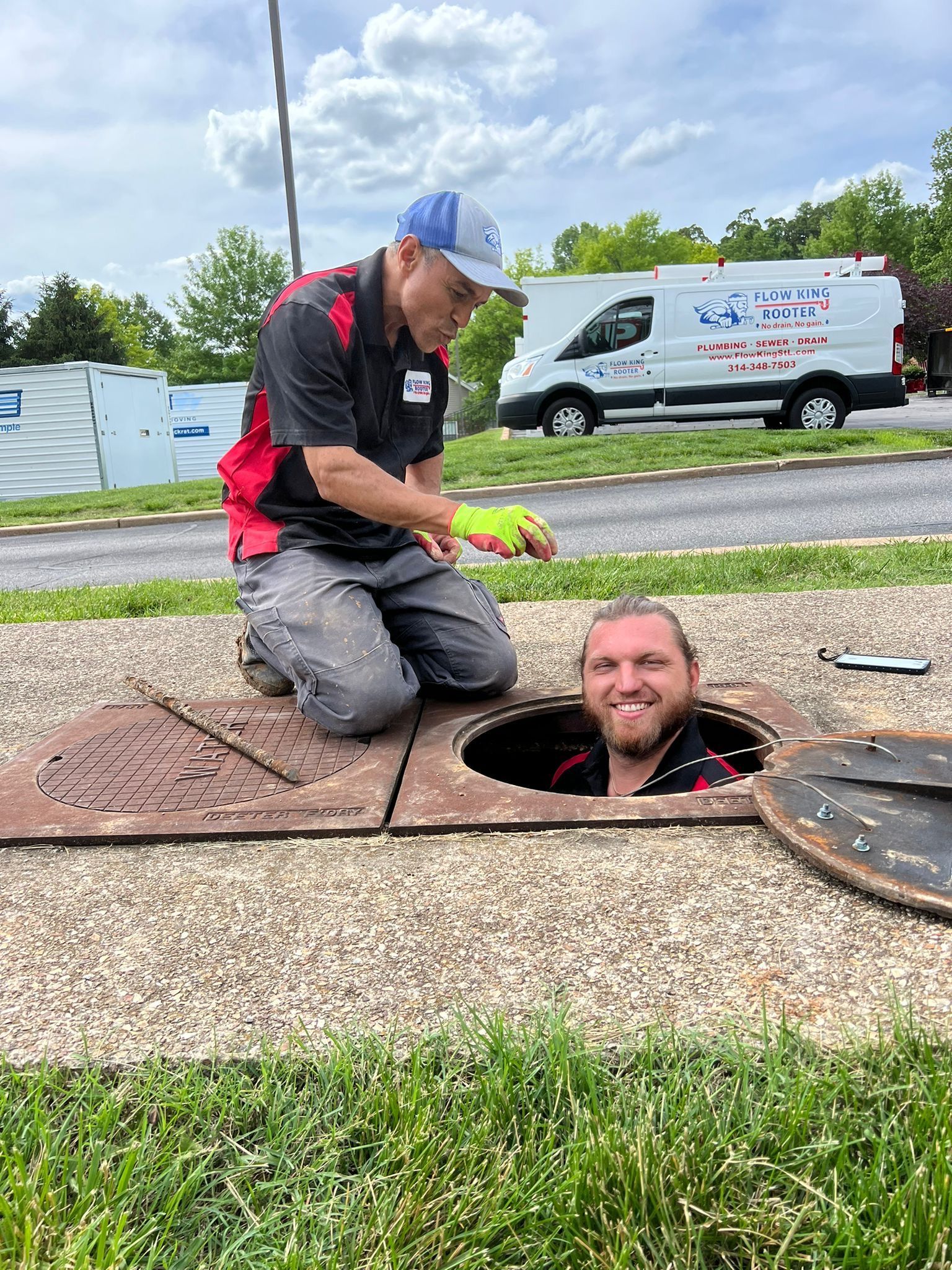 Two men working on an open manhole in a grassy area with a van in the background. One man is inside the manhole.