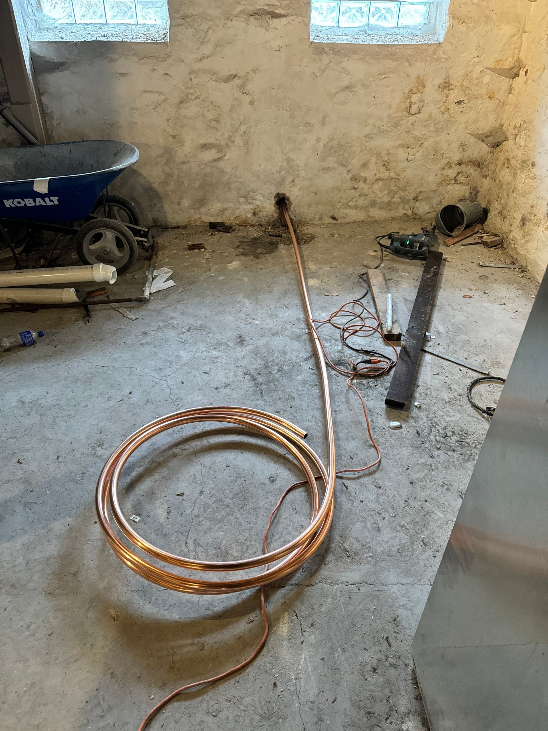 Copper tubing coiled on a concrete floor in a basement with tools and a wheelbarrow.