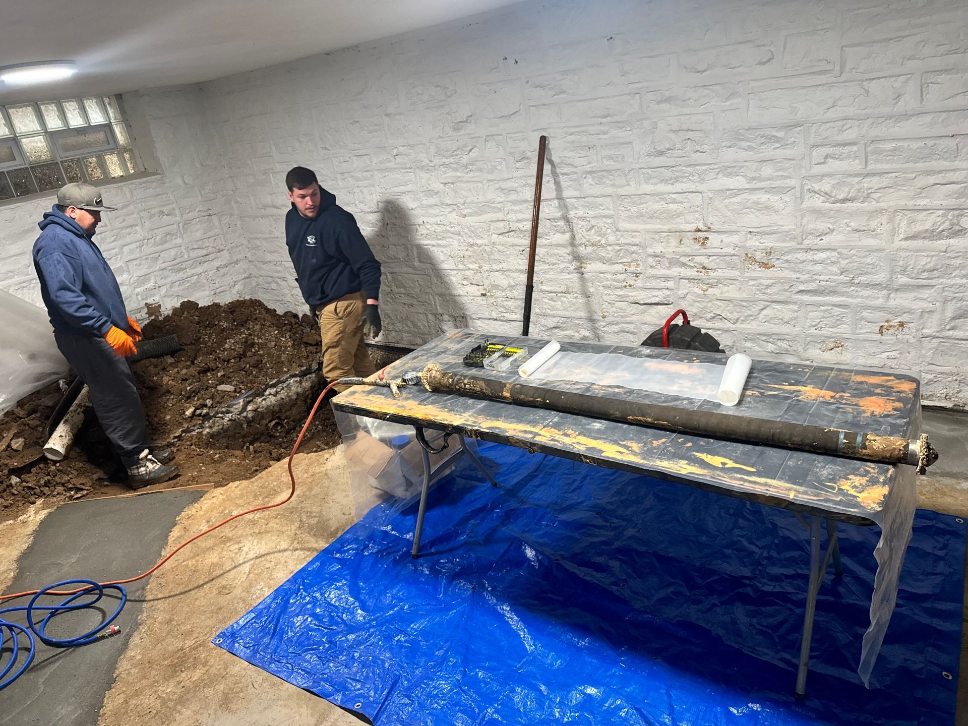 Two people working in a basement. One shovels dirt; the other stands nearby. A table covered in plastic is in the foreground.