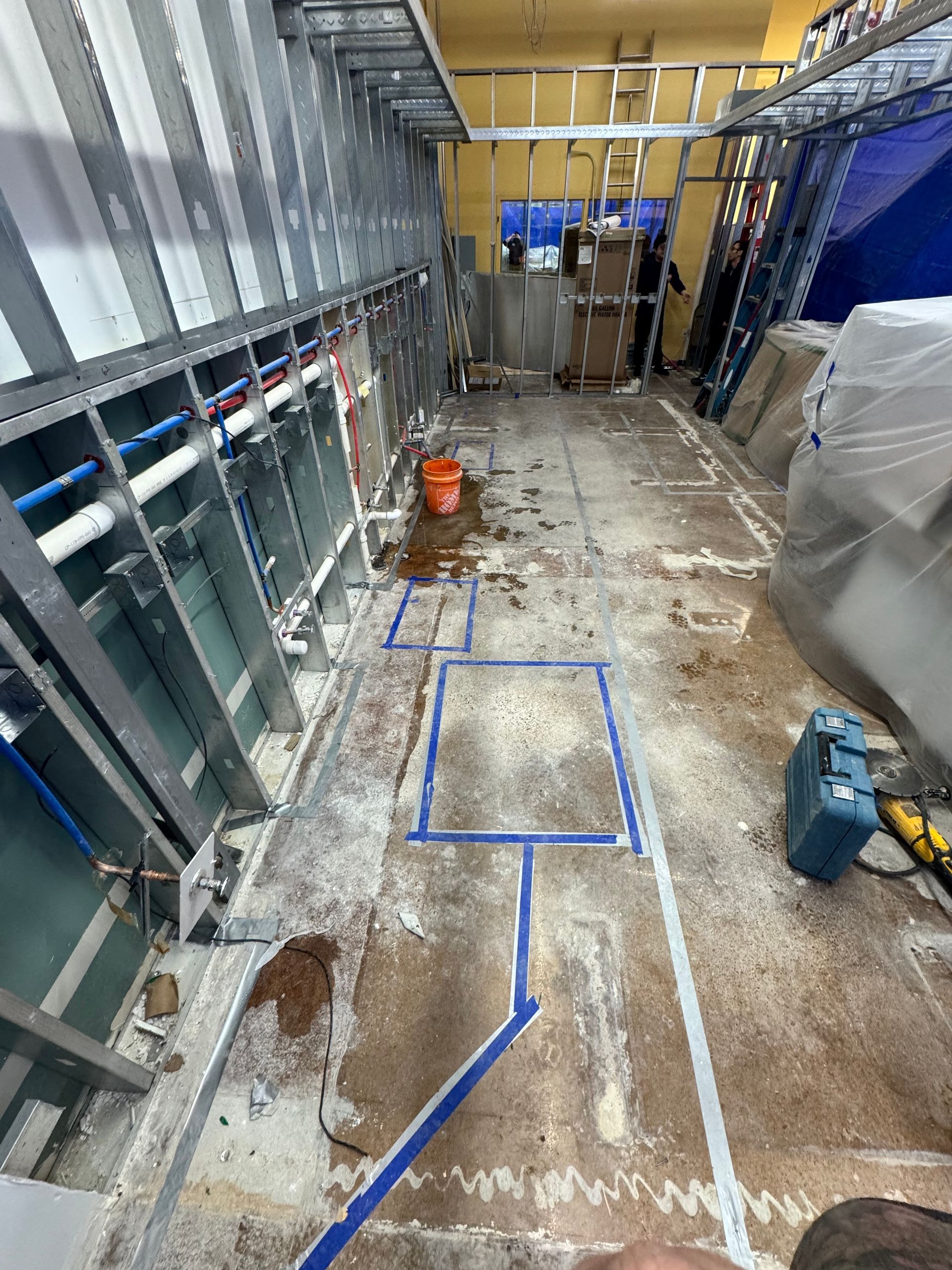 Construction site with metal framing, blue plumbing, and marked floor plans. A bucket and tools are visible.