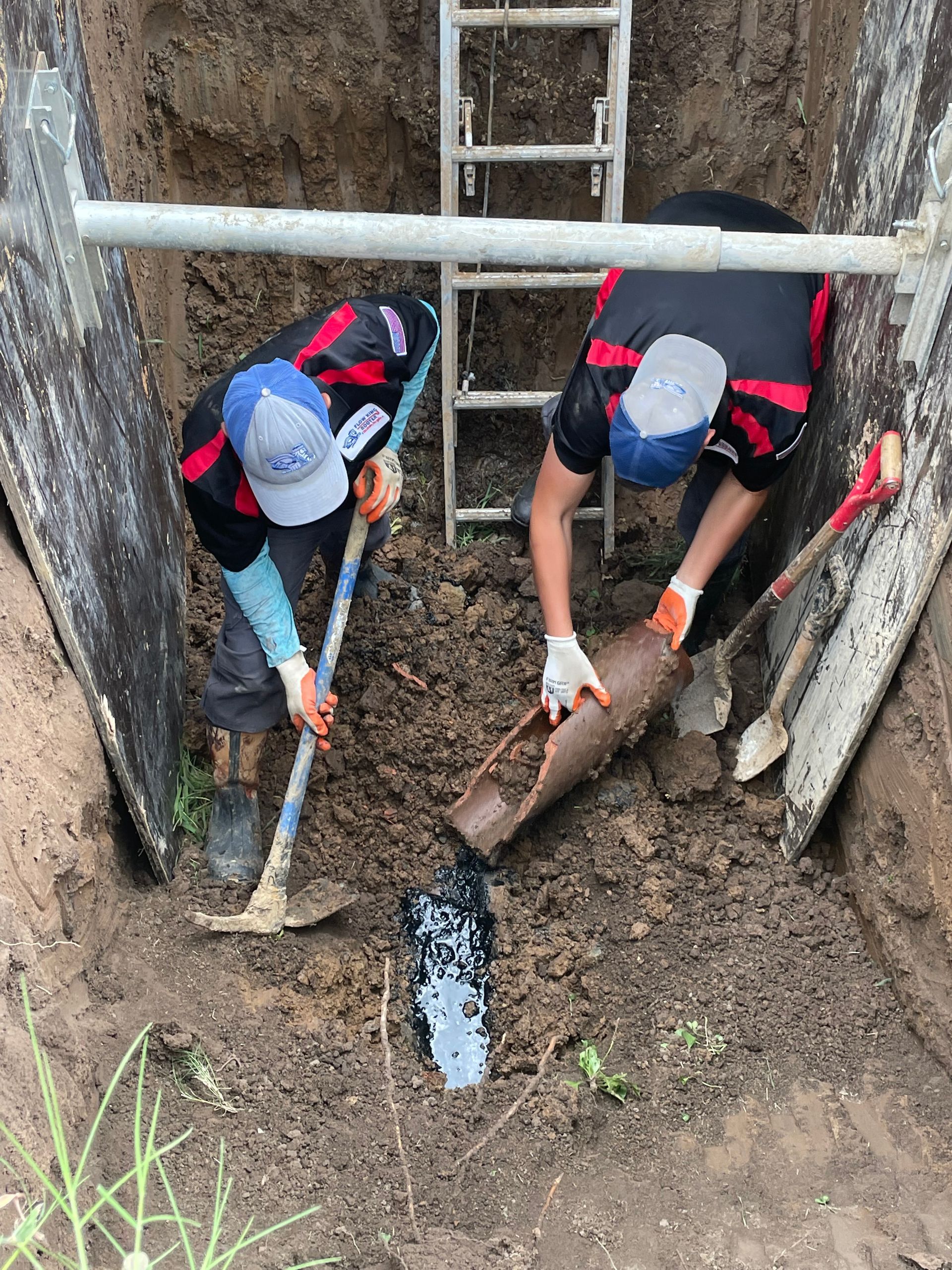 Two workers in a trench fix a broken pipe, using shovels. A ladder and support beam are visible.