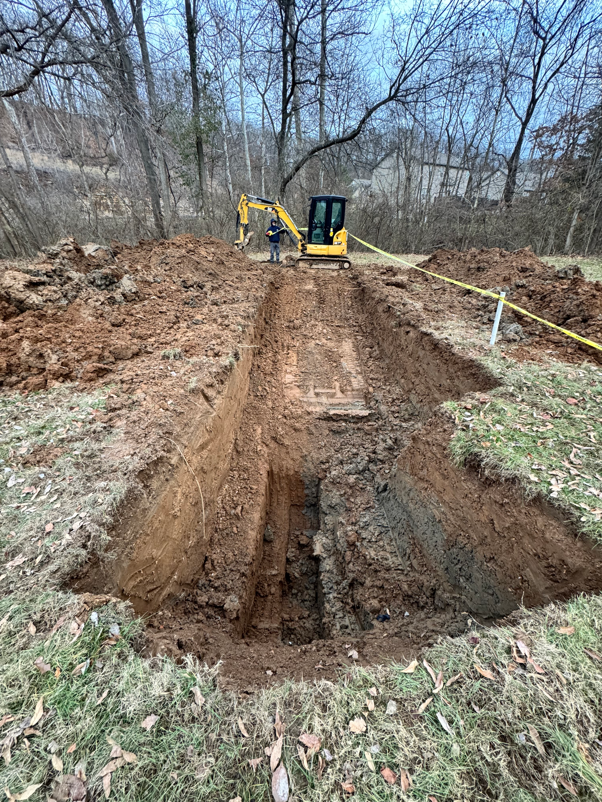 Excavator digging a trench in a grassy area, revealing a buried structure. Trees in the background.
