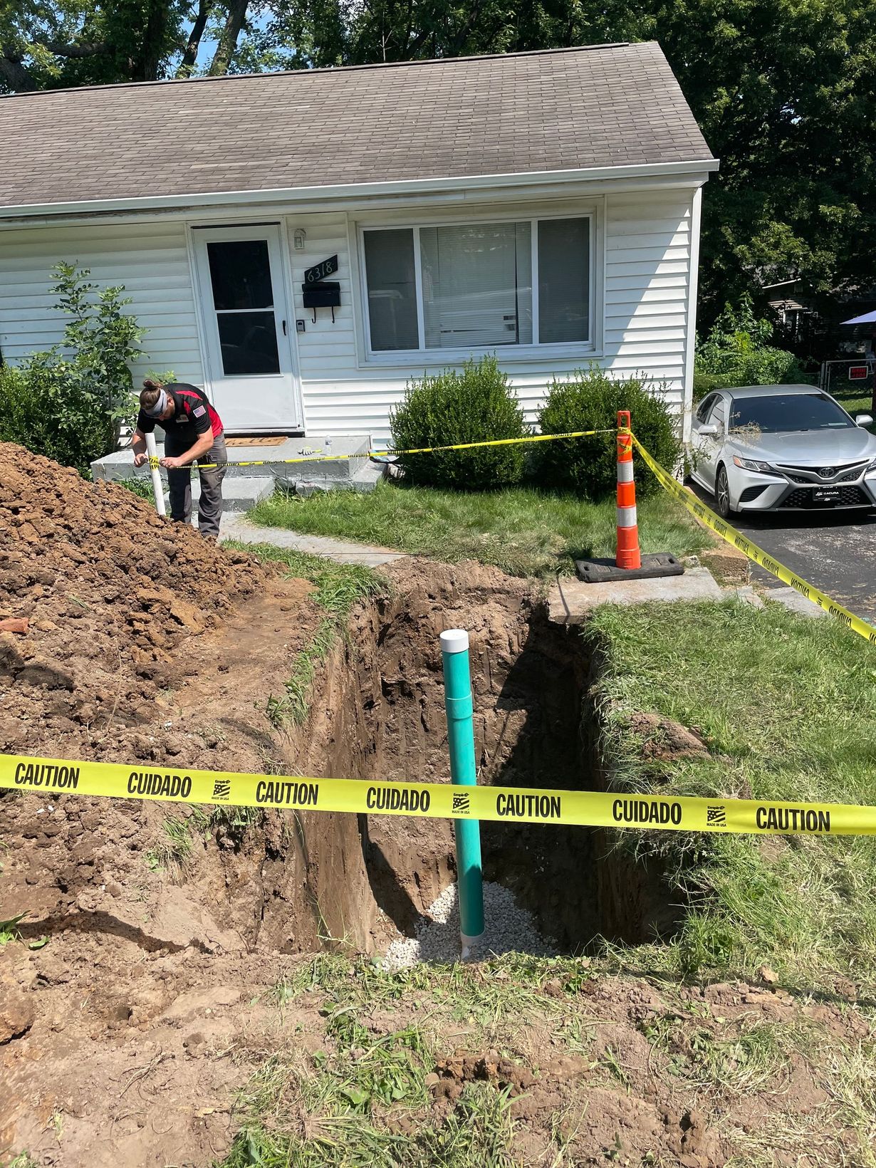 Man near an open trench at a house. Yellow caution tape surrounds the excavation, containing a green pipe.