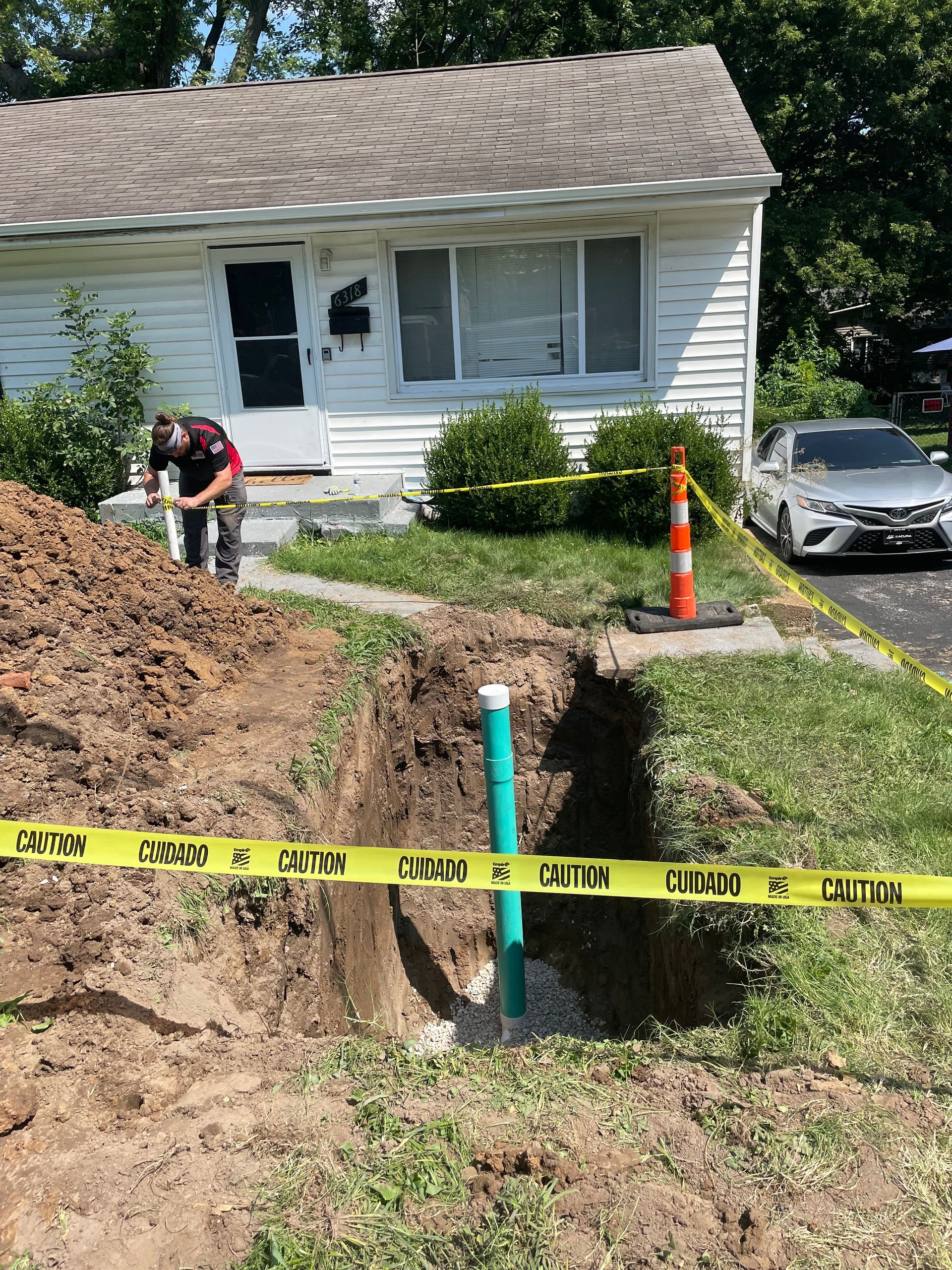 Man near an open trench at a house. Yellow caution tape surrounds the excavation, containing a green pipe.