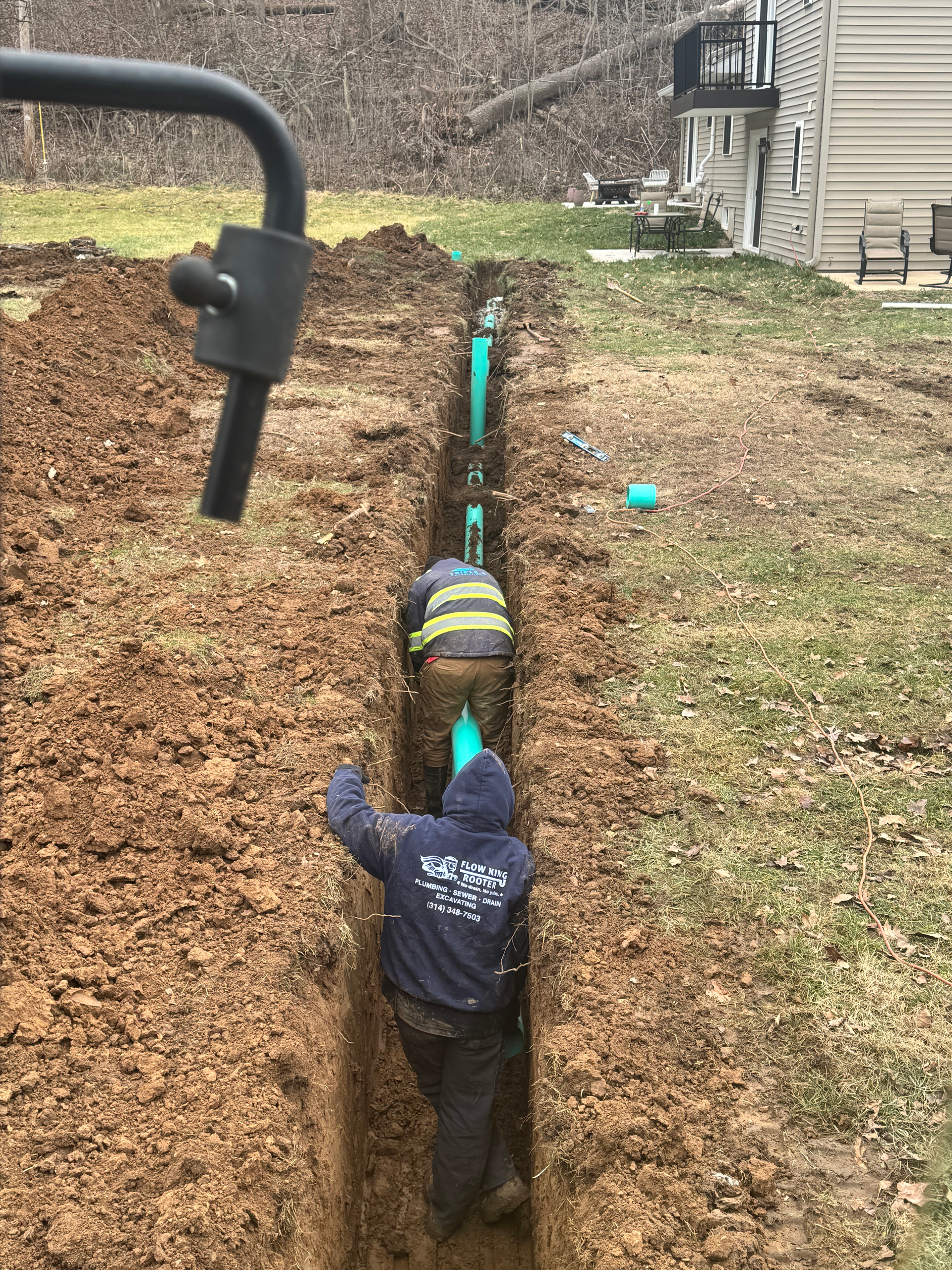 Two people install green pipes in a narrow trench in a grassy yard.