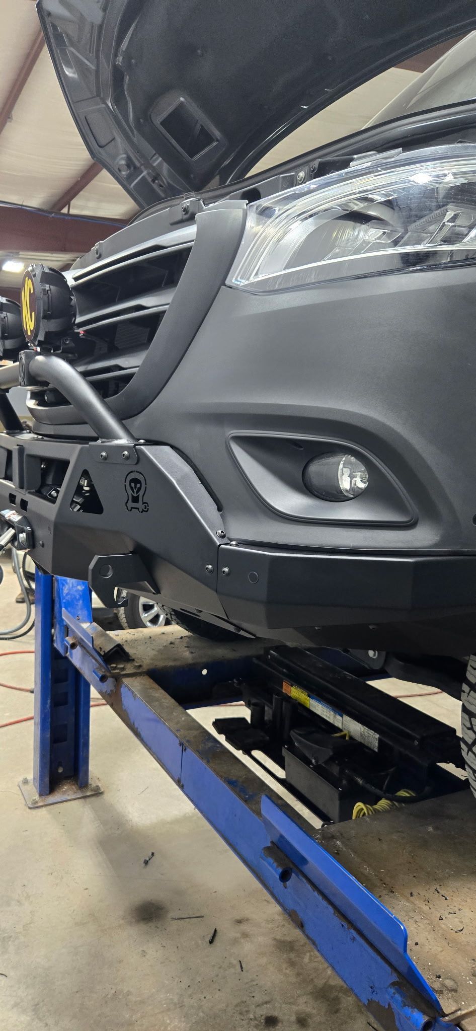 Black van on a blue car lift, its front bumper replaced with a black, custom-made one.