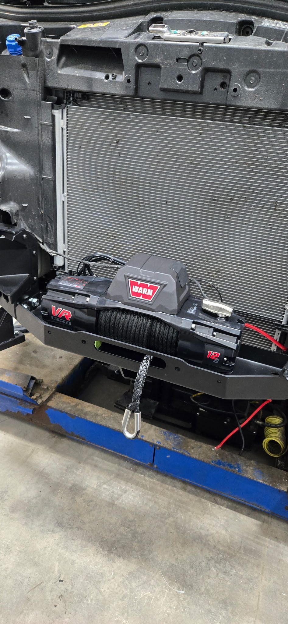 Black vehicle bumper with a winch. A radiator is behind the bumper, and the vehicle is on a blue lift.