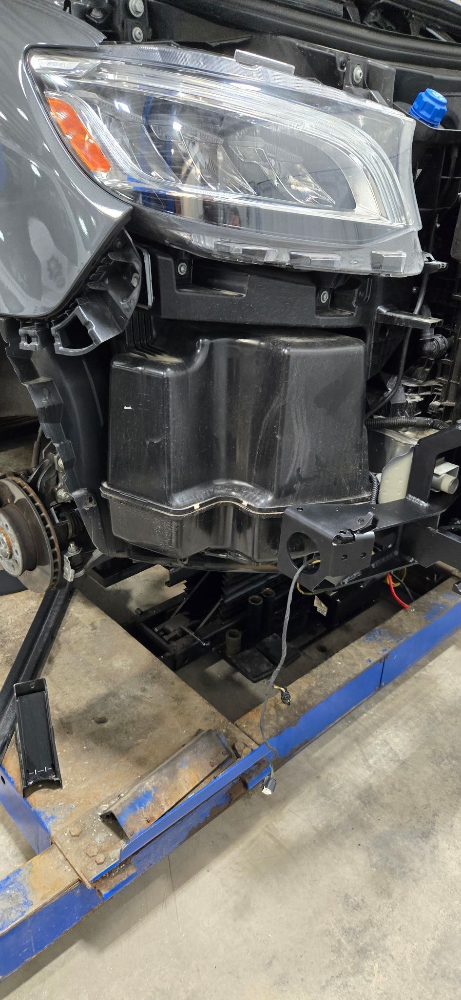 Front of a gray car's frame, headlight visible. A black component sits between the headlights.