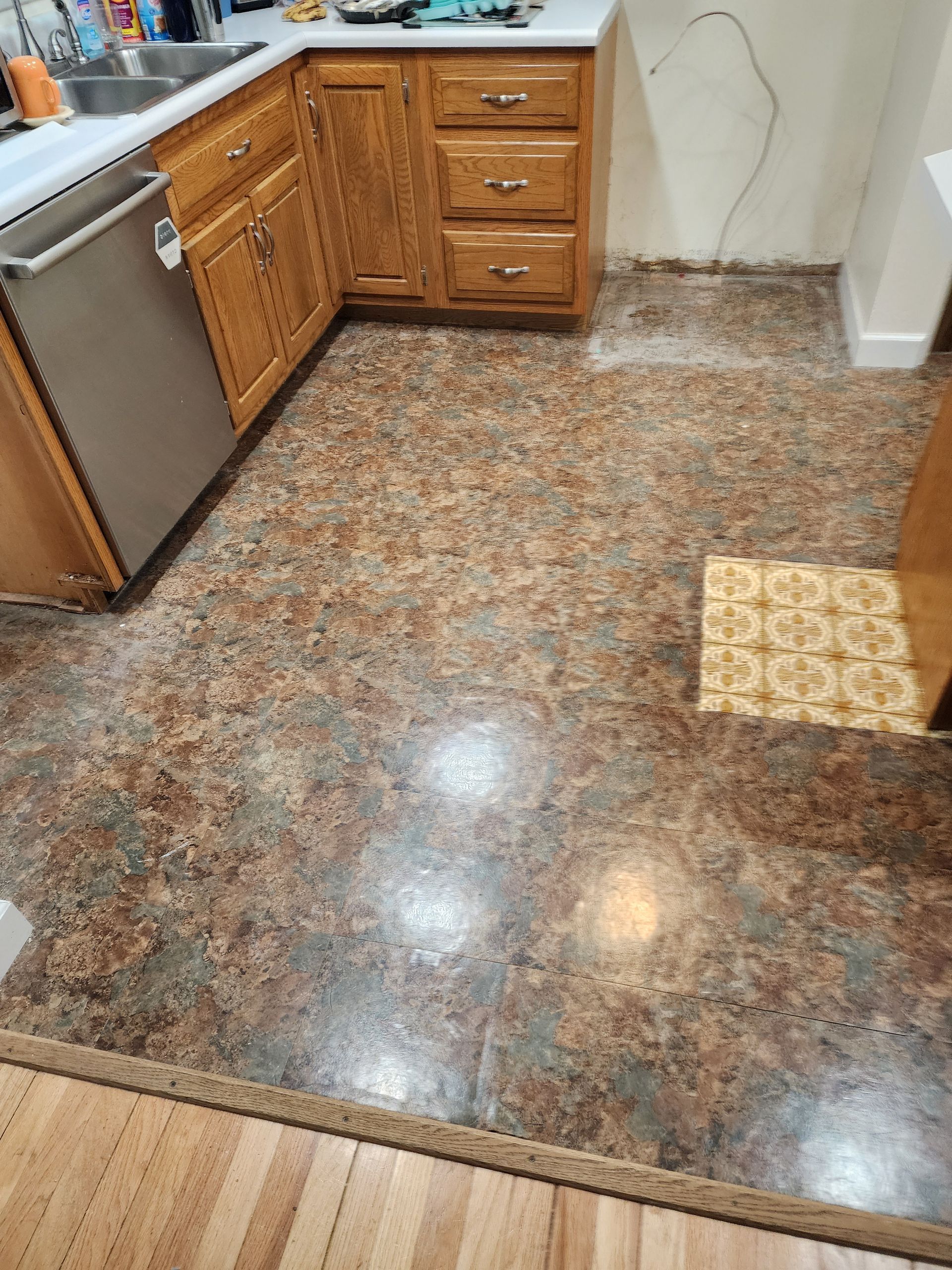 Kitchen with speckled brown floor, wood cabinets, and a stainless dishwasher by a white wall.