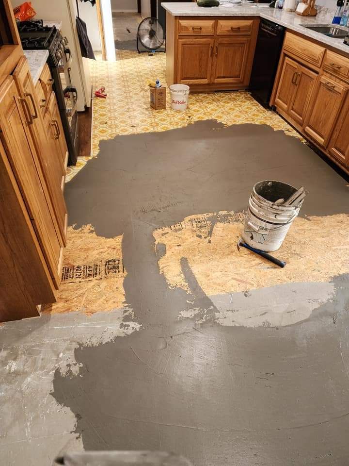 Kitchen floor mid-renovation with gray patching compound spread over removed tile and a bucket nearby